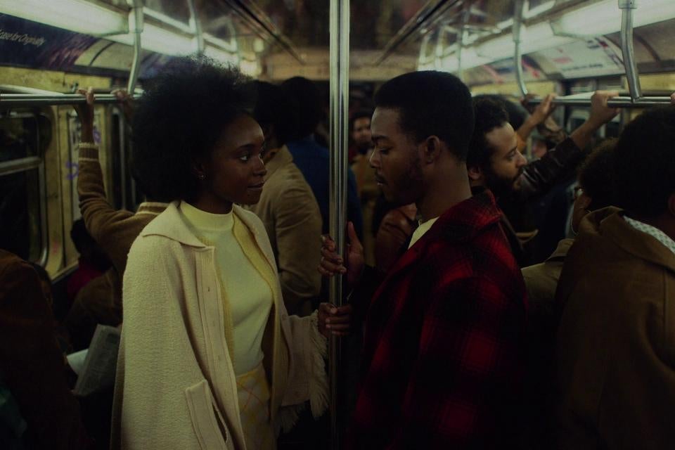 In the 1970s, a young Black woman and young Black man stand, staring intently at each other, on a crowded subway car while both holding onto the pole in the middle of the car.