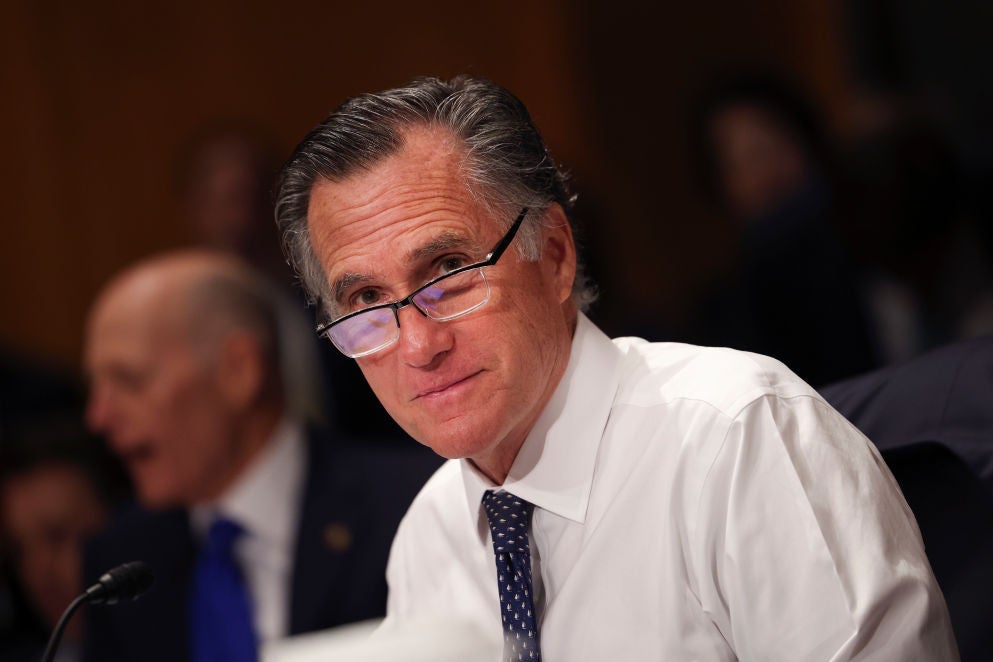 Mitt Romney, seated, in shirtsleeves, tie, and glasses, looks toward the camera and smiles