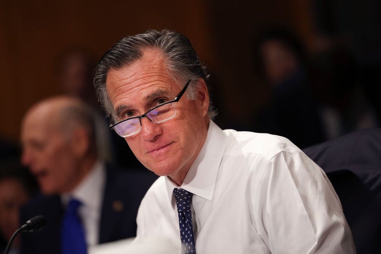 Mitt Romney, seated, in shirtsleeves, tie, and glasses, looks toward the camera and smiles