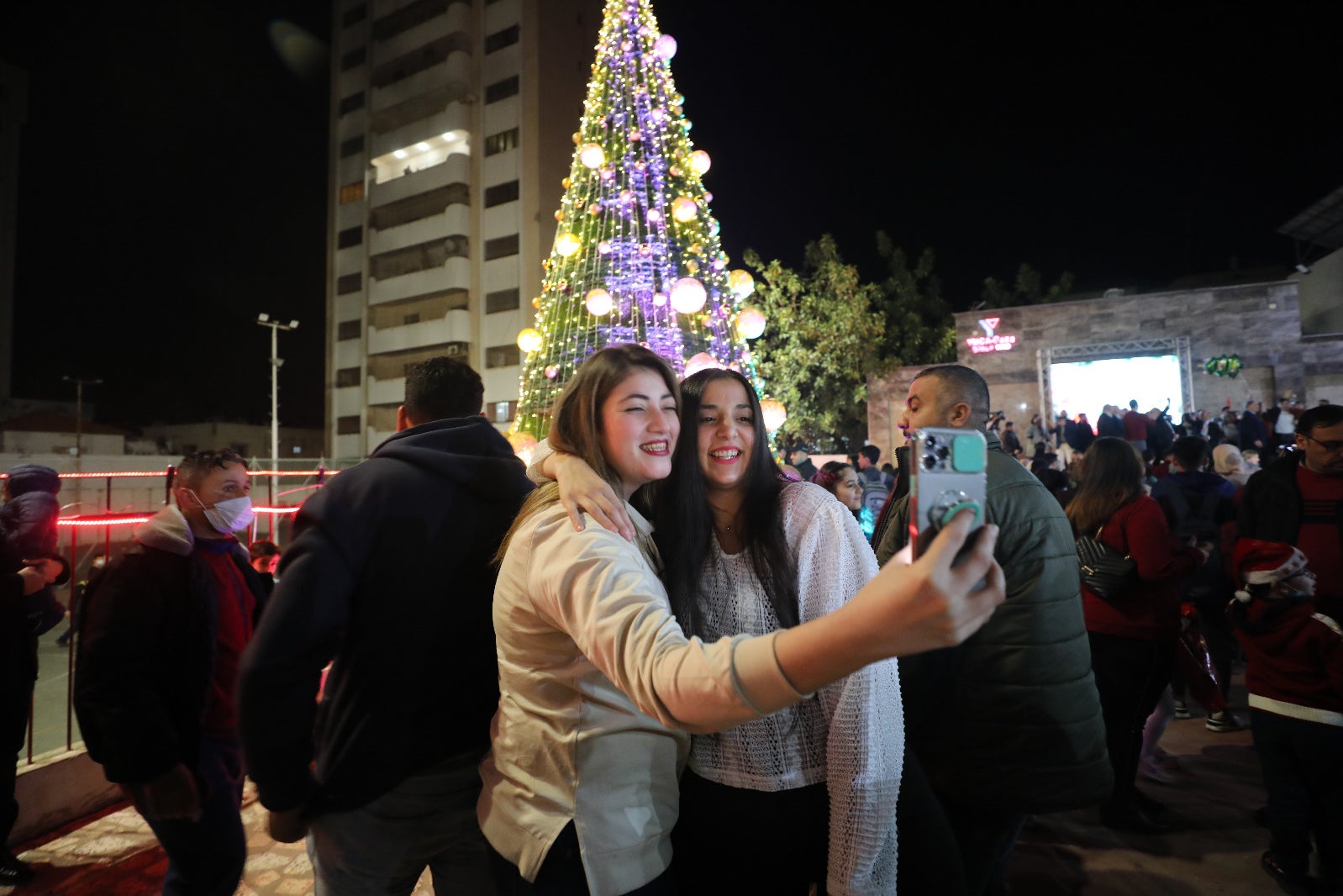 Two teenage girls take a selfie in front of a Christmas tree bedecked with lights in ornaments, smiling and embracing.