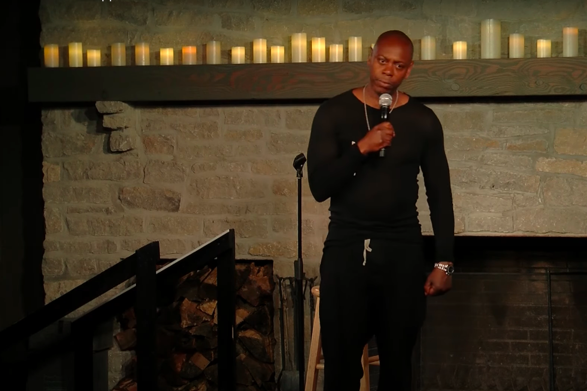 Dave Chappelle standing, one hand holding his microphone and the other clenched in a fist
