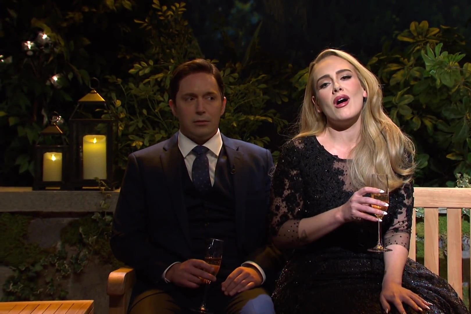 Beck Bennett and Adele sit on a wooden bench on a candlelit patio. Adele is singing while Bennett gives her a side-eyed look.