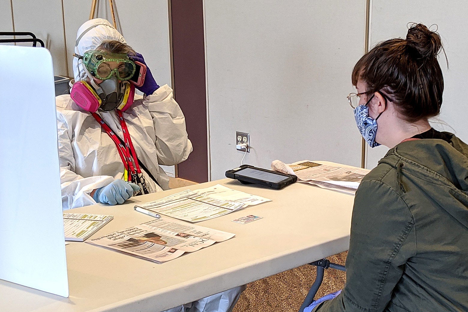 A woman in a hazmat suit sits across a table from a woman in a face mask at a polling station.