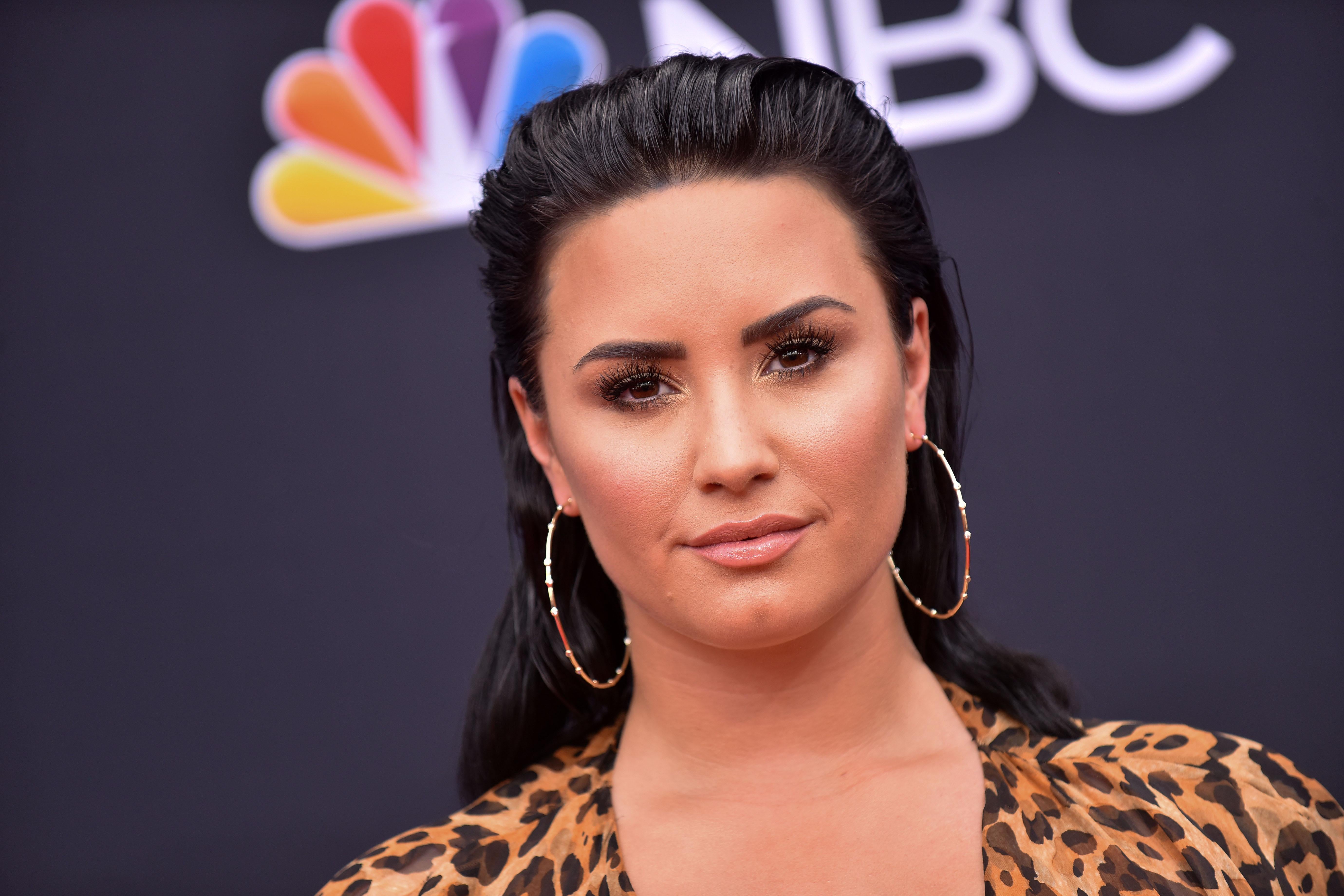 Demi Lovato on a red carpet in front of the NBC logo. She wears hoop earrings and leopard print.