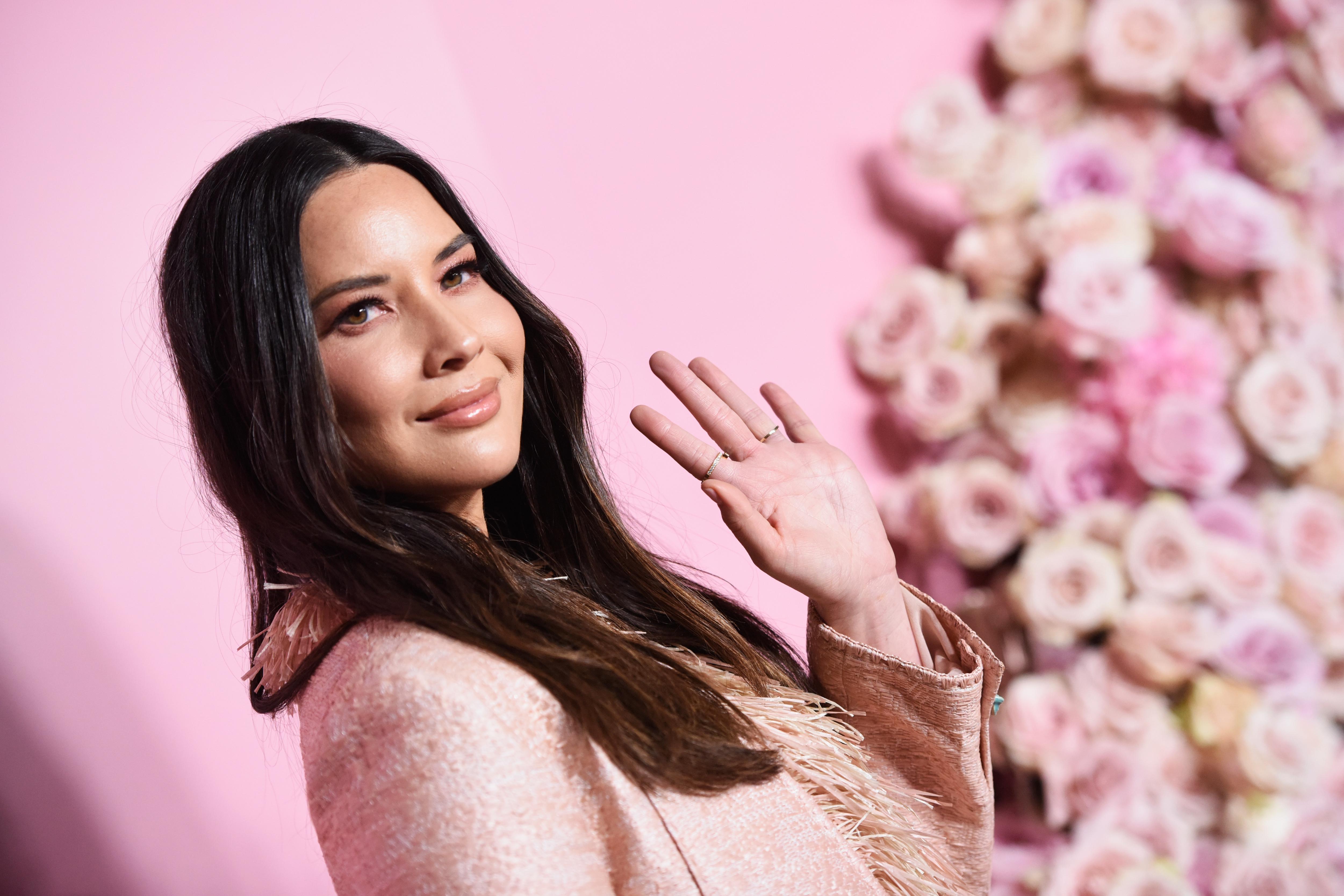Olivia Munn from the chest up, waving, in a skewed-frame picture with pink background.