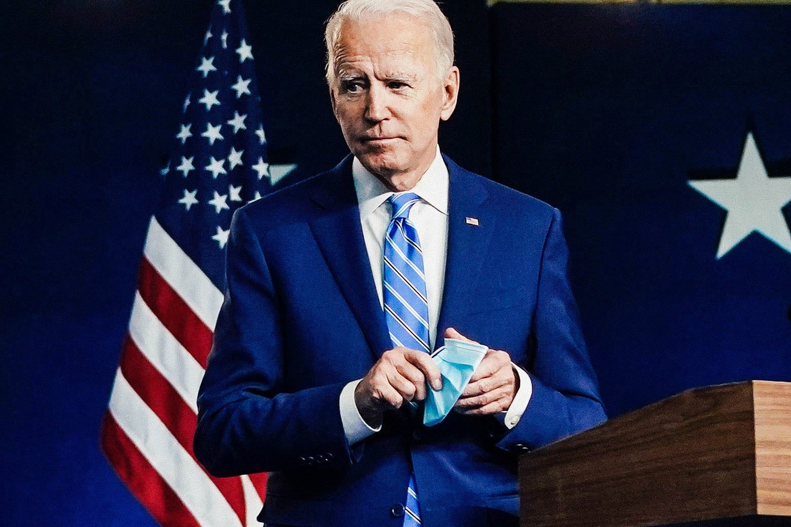 Biden, holding a surgical mask, walks away from a lectern, with an American flag in the background