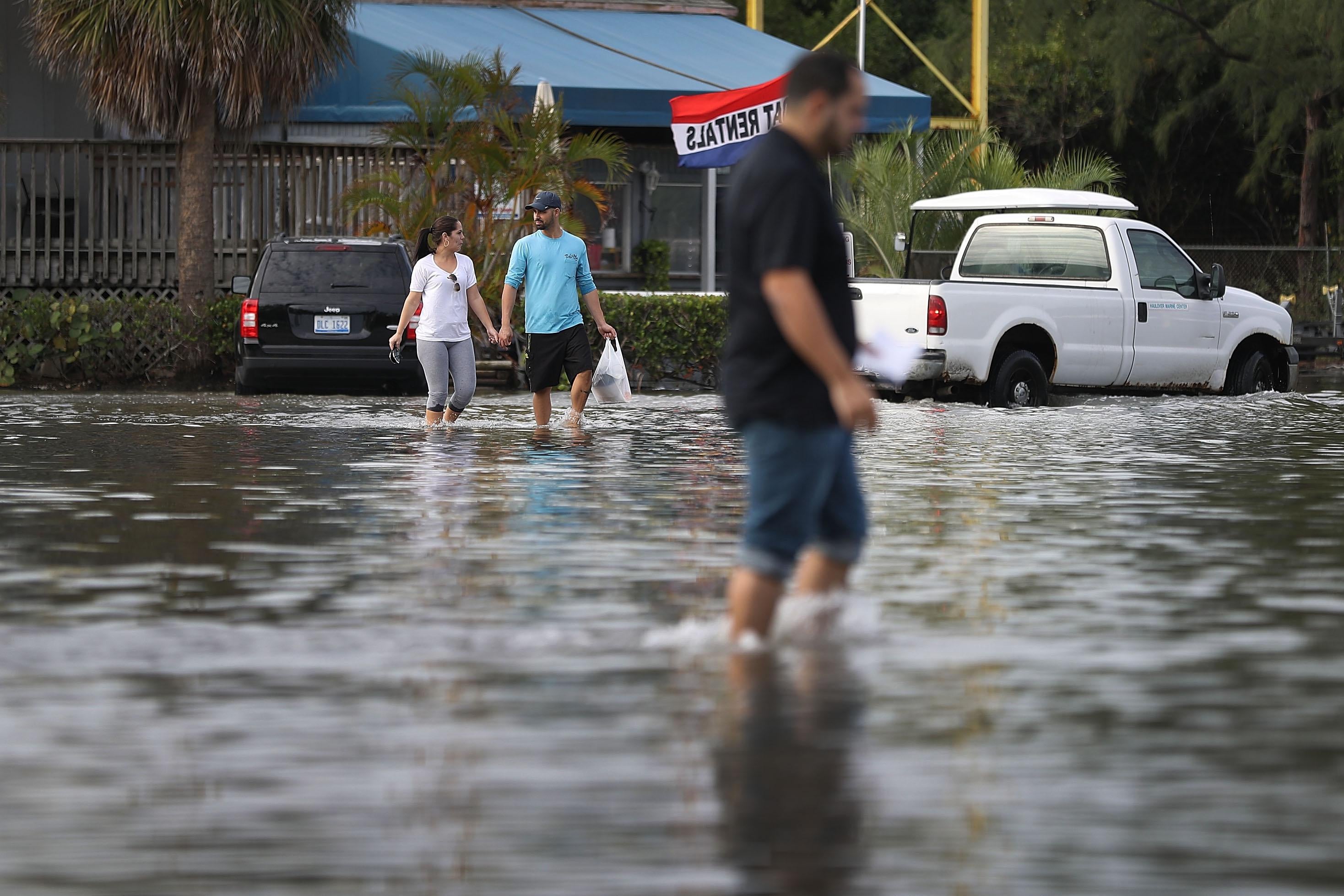 NORTH MIAMI, FL - NOVEMBER 14:  Yaneisy Duenas (L) and Ferando Sanudo walk through the flooded parking lot to their boat at the Haulover Marine Center on November 14, 2016 in North Miami, Florida. The flood waters are caused by the combination of the lunar orbit which causes seasonal high tides, also known as a King tide, and what some scientists believe is rising sea levels due to climate change.  (Photo by Joe Raedle/Getty Images)