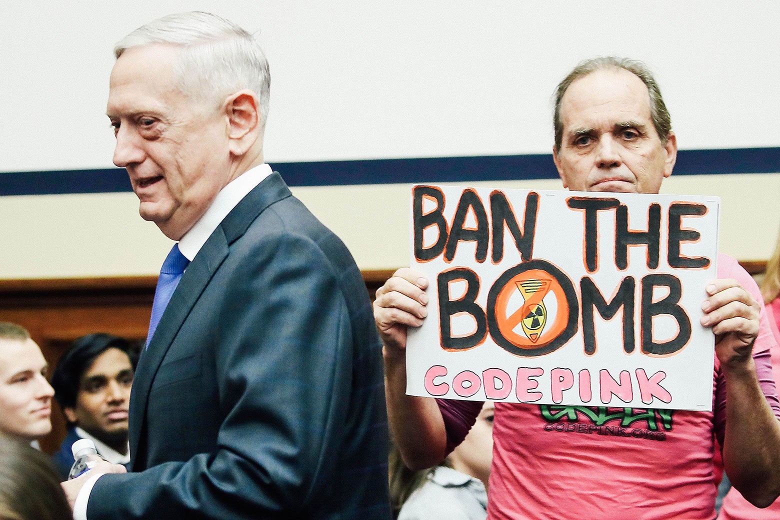 Defense Secretary Jim Mattis walks past a protester holding a sign that says, "BAN THE BOMB. CODE PINK."