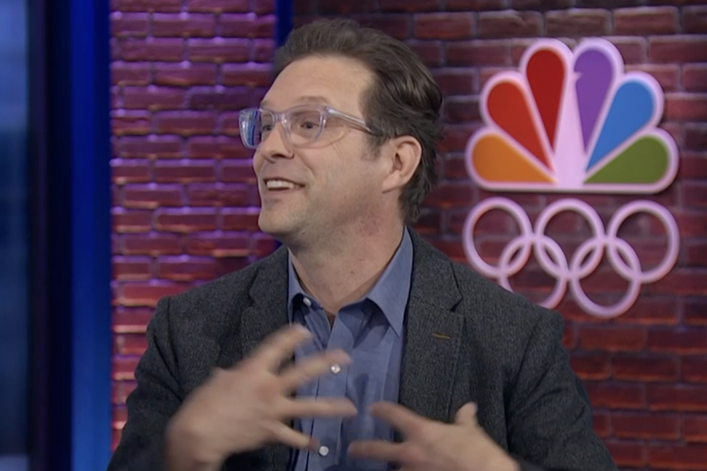 Chad Salmela speaking in studio gesturing at himself with NBC's Olympics logo over his shoulder.