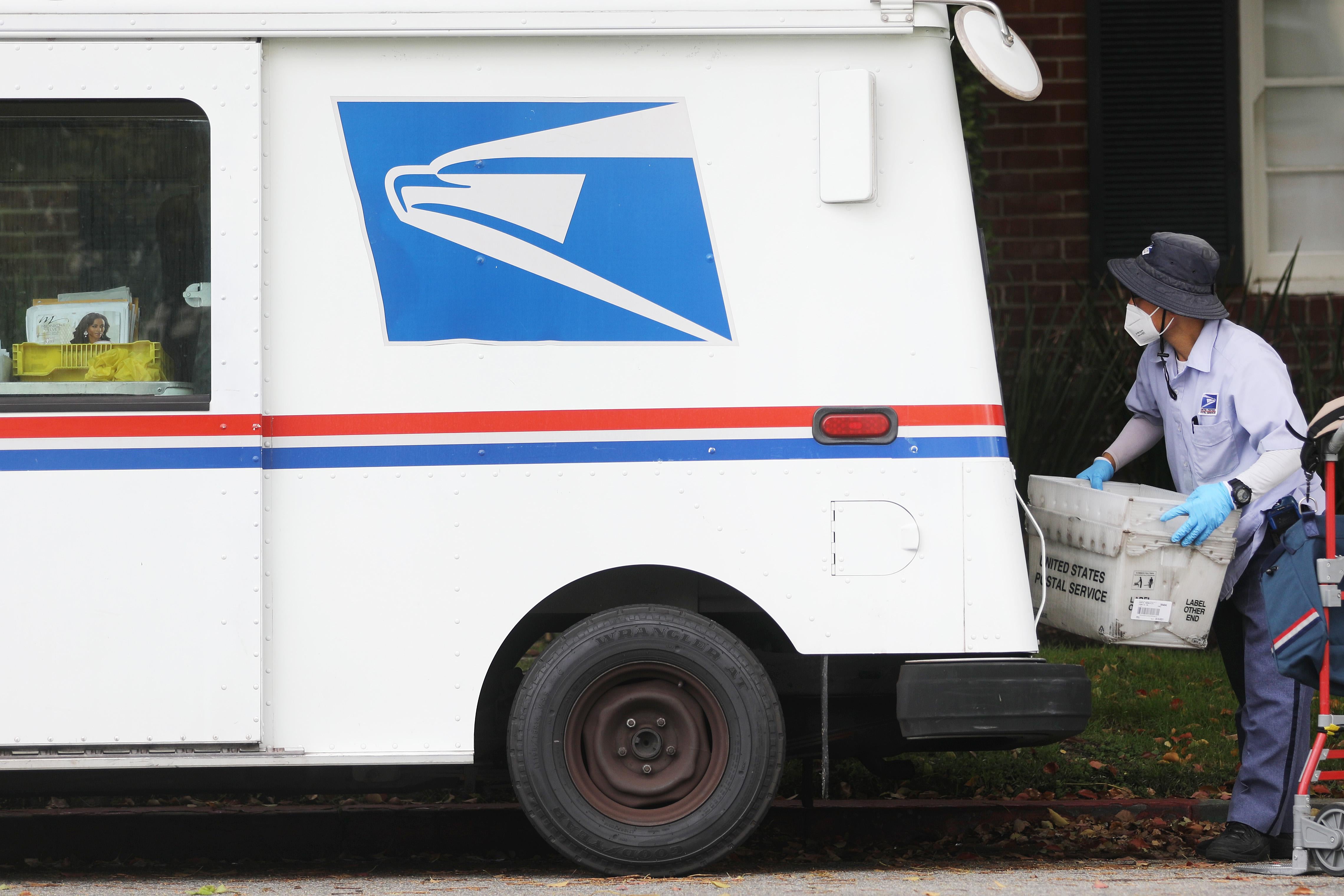 A postal worker wears gloves and a mask while carrying a mail bin near a truck.