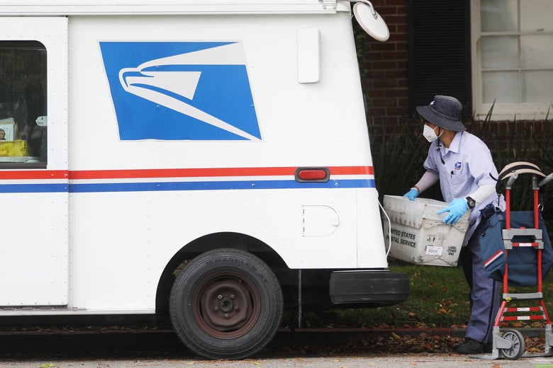 A postal worker wears gloves and a mask while carrying a mail bin near a truck.