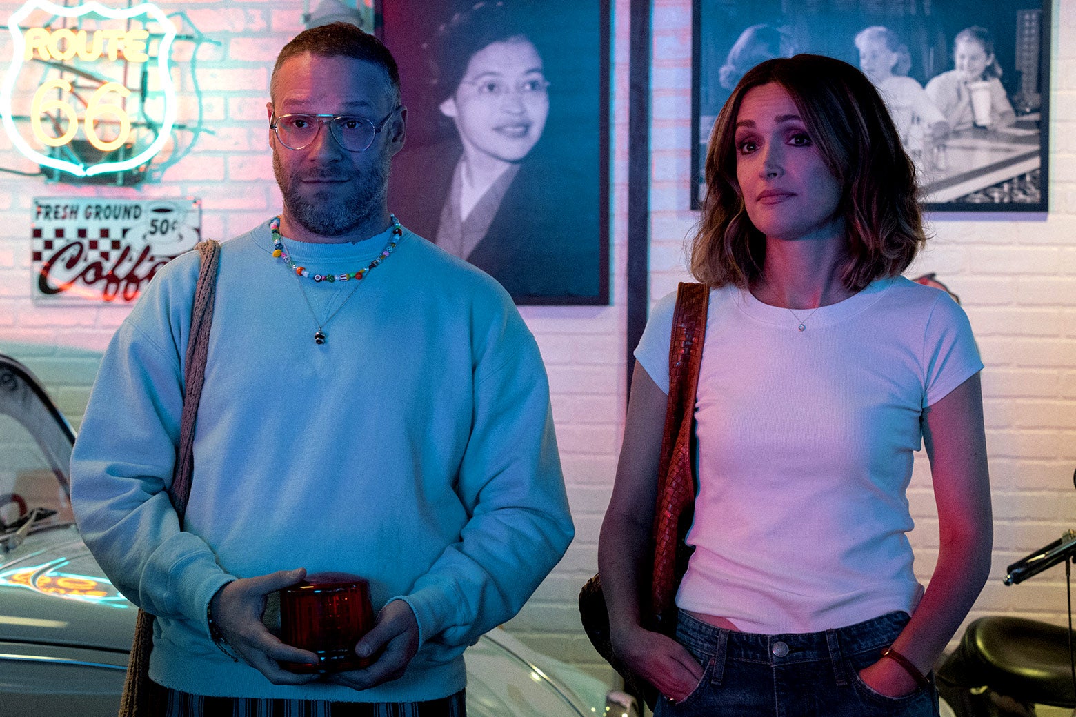 A middle-aged white man in glasses, who's wearing a plain blue sweatshirt with a child-created rainbow-bead necklace, stands next to a middle-aged white woman wearing a white T-shirt as both stare ahead. Neither looks impressed.