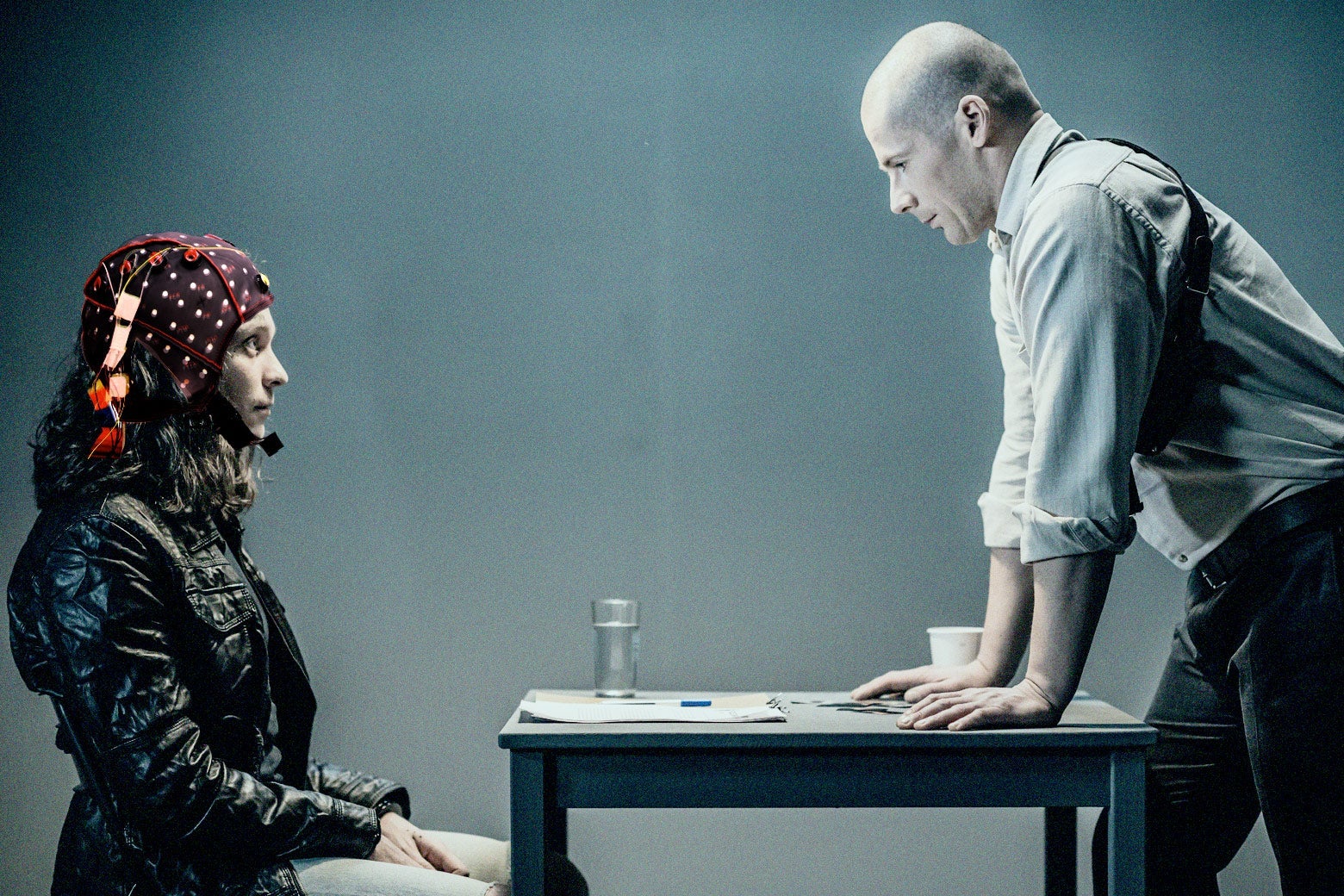 A woman sits in front of a table wearing a helmet with electrodes, while a man stands on the other side, his hands on the table, looming.