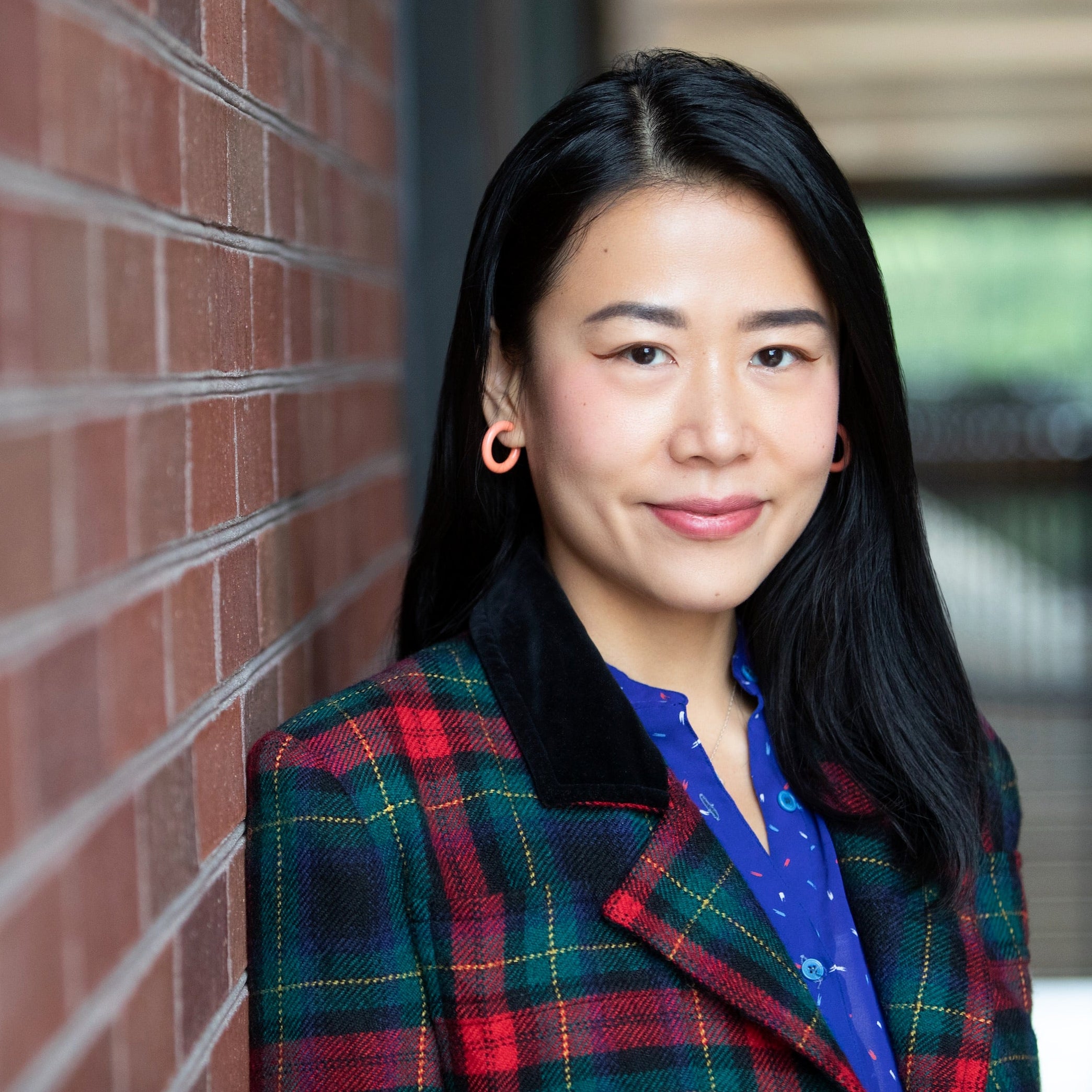 A woman with black hair stands next to a brick wall.