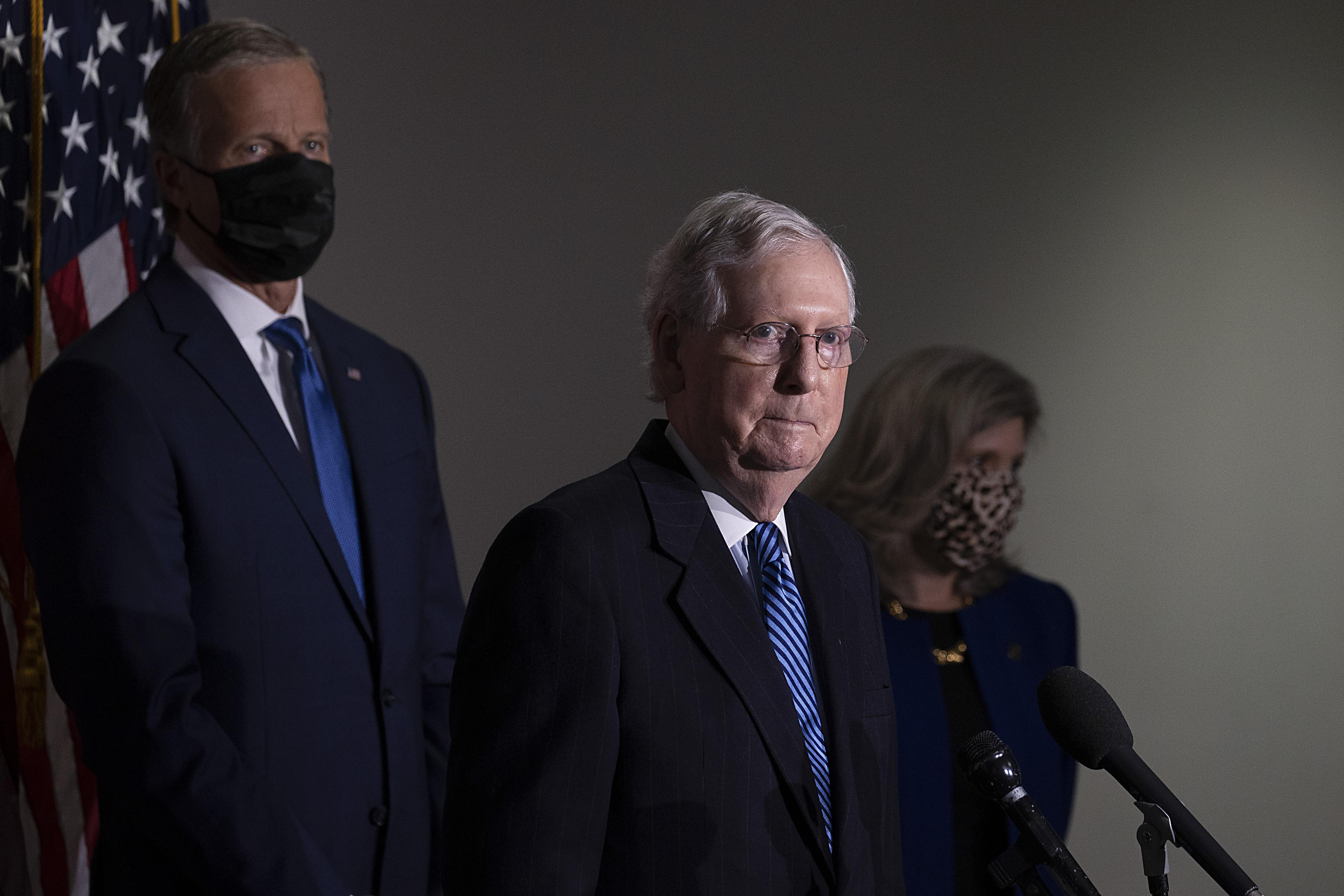 Mitch McConnell speaks at a mic with two people flanking him