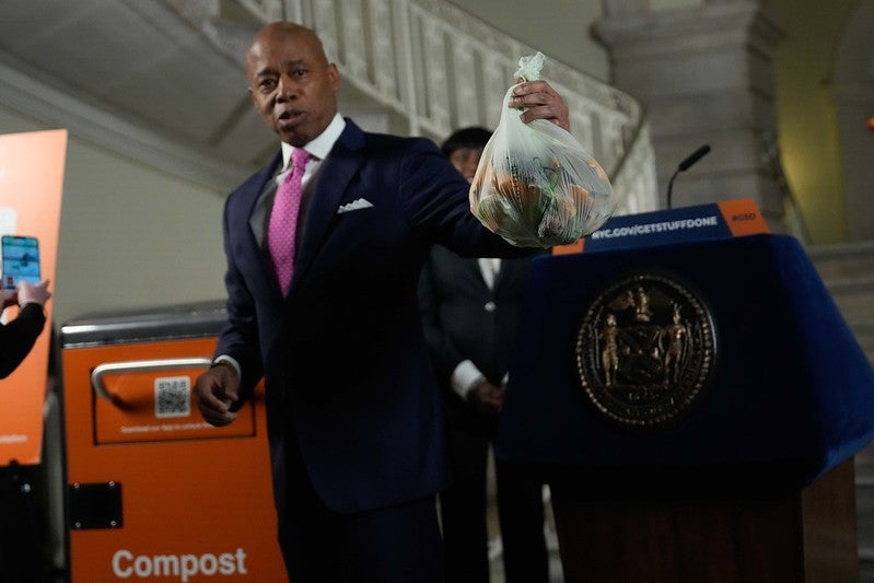 Eric Adams holds up a bag full of compost while standing in front of a lectern and a composting bin.