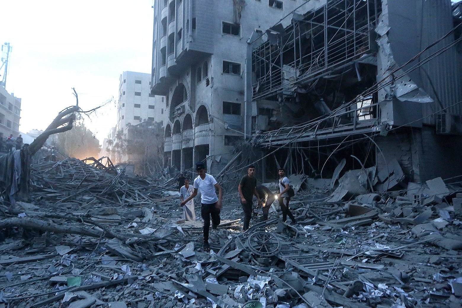 A group of four Palestinians step across rubble. Behind them is a bombed-out building and a tree that has been flipped upside-down.