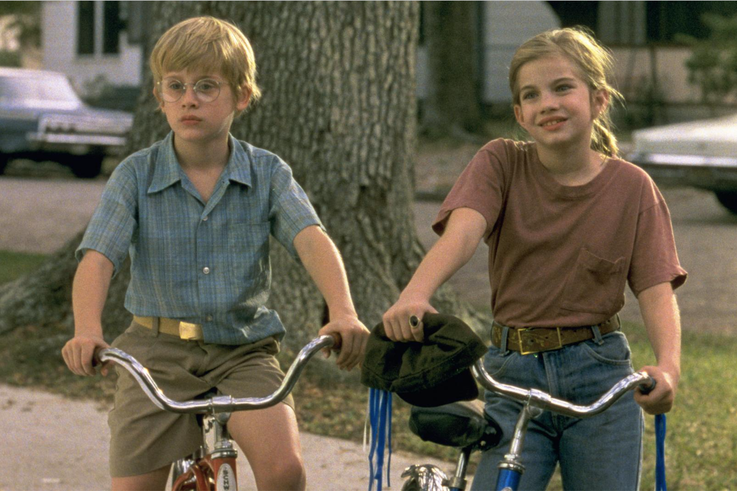 Two young children pause on the sidewalk on their bikes: on the left a young blonde boy with glasses and a patterned button down (a young Macaulay Culkin), on the right a young blonde girl in a pony tail in a t-shirt and jeans (a young Anna Chlumsky).