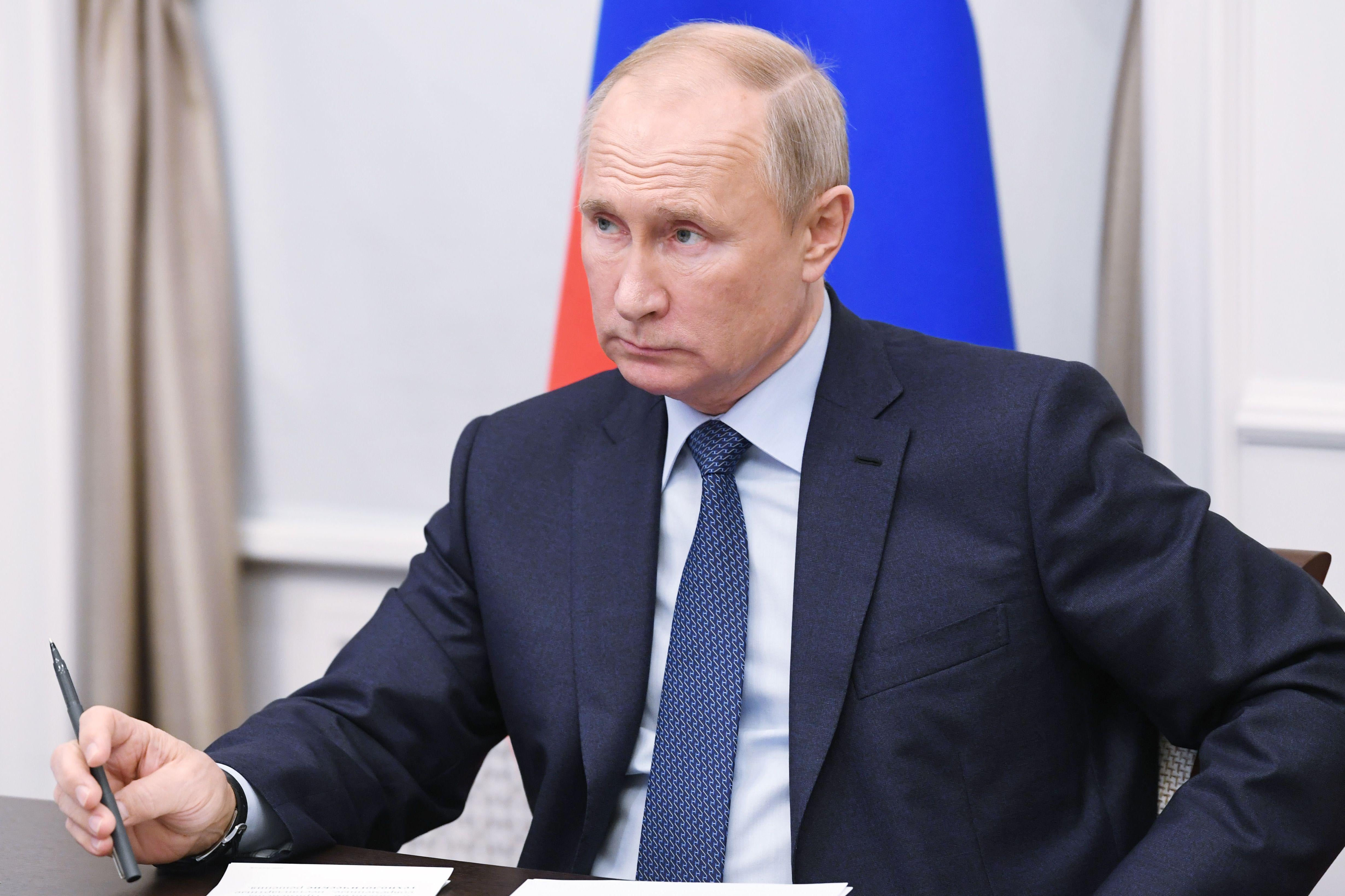 Putin sitting at a desk holding a pen, with a Russian flag behind him