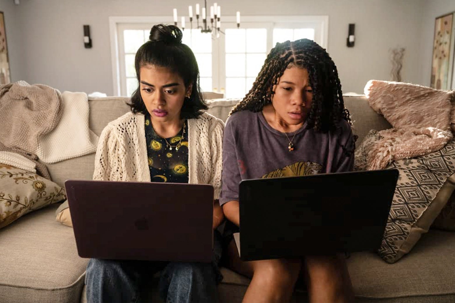 Two girls from the movie sit next to each other on a couch with laptops open and scared looks on their faces.