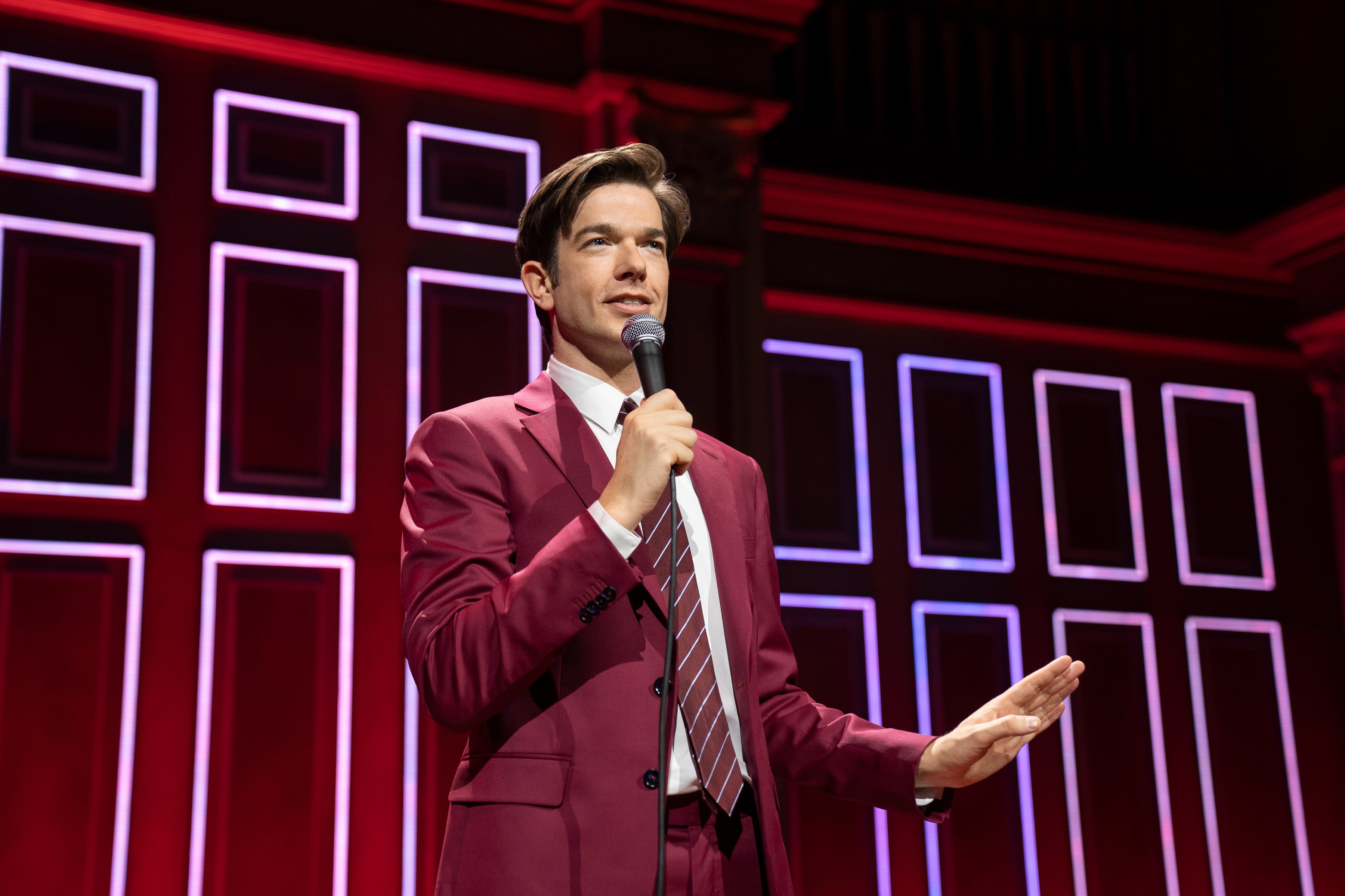 He stands on stage in a burgundy suit, his hair floppy. In his right hand, he holds a microphone. In his left, he holds up a hand, as if to say "wait a minute.“