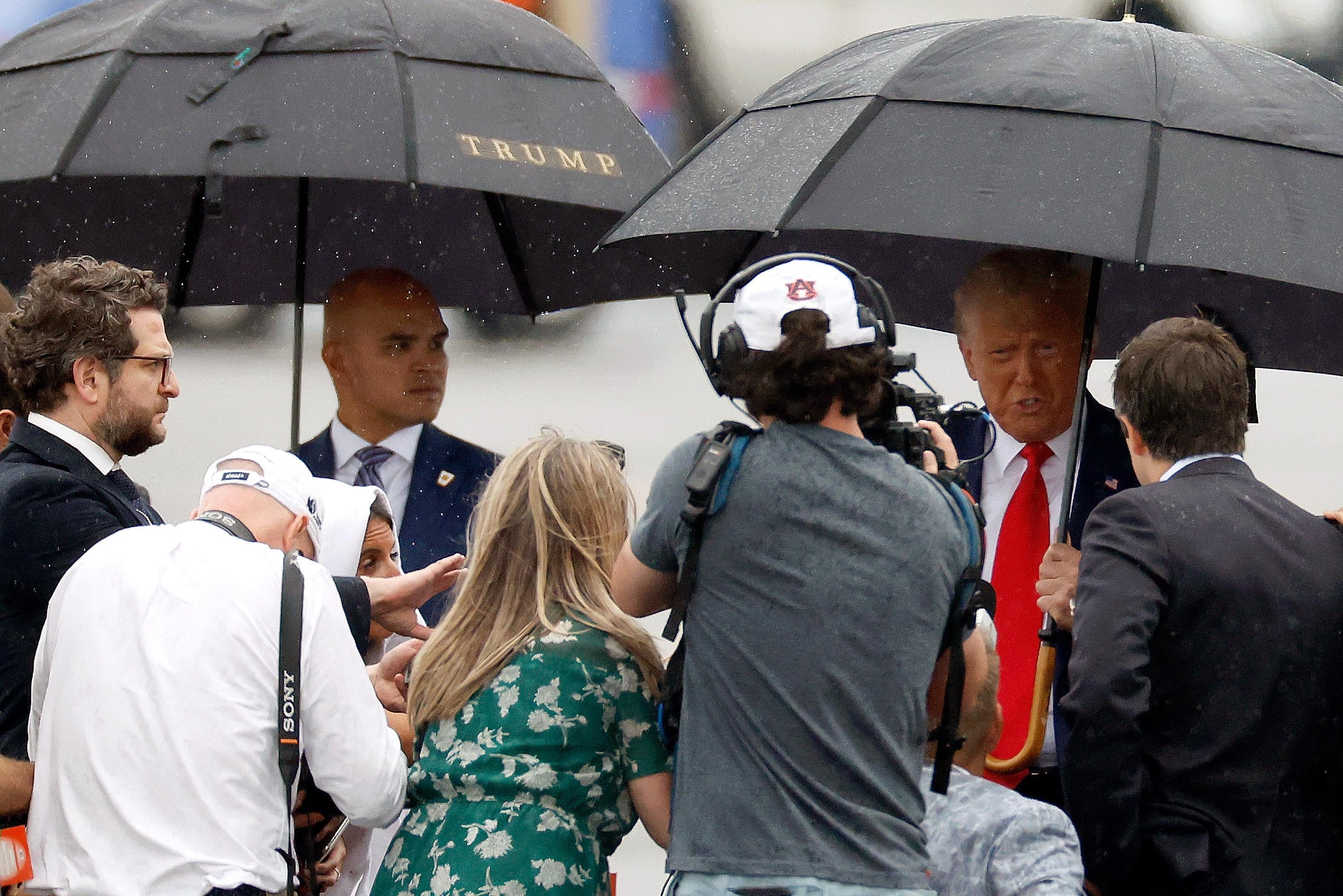 A man holding an umbrella speaks to a crowd of media, while a body guard with another umbrella stands by.