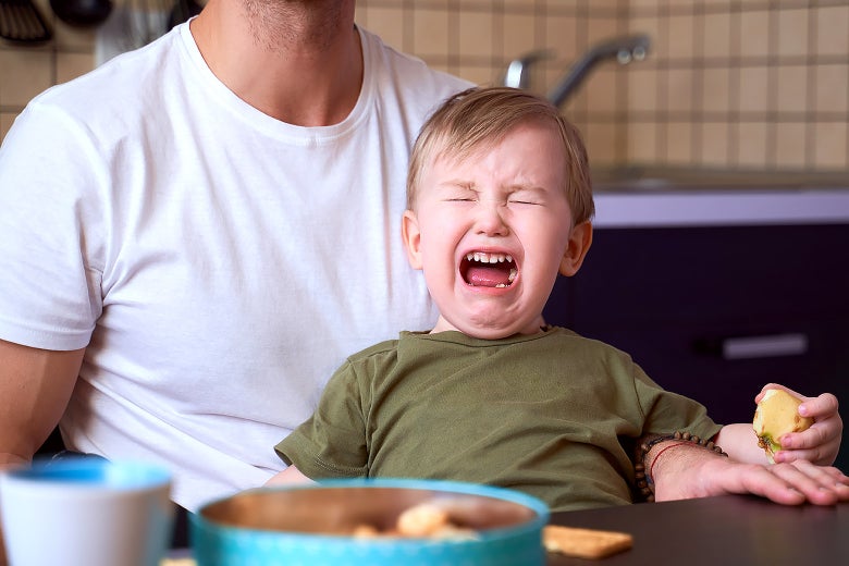 A young boy having a meltdown in his dad's lap at the kitchen table.