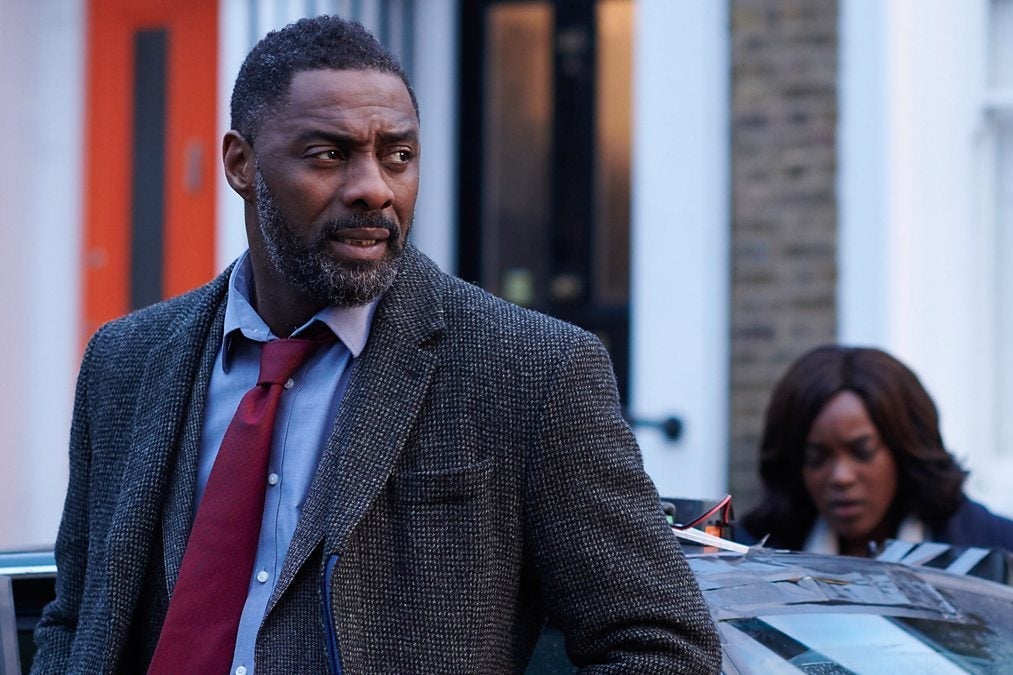 Idris Elba stands in front of a car. Wunmi Mosaku, blurry, in the background.