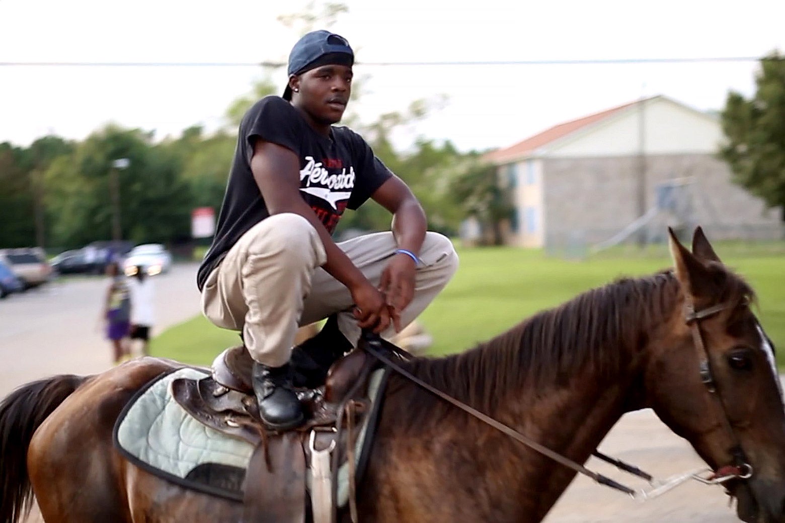 A young man in a T-shirt and khakis crouches on a horse.