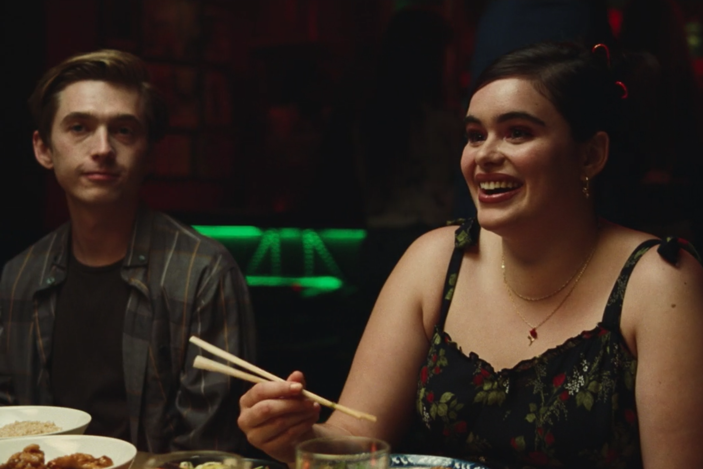 A girl with black hair sits at a table holding chopsticks and smiles.