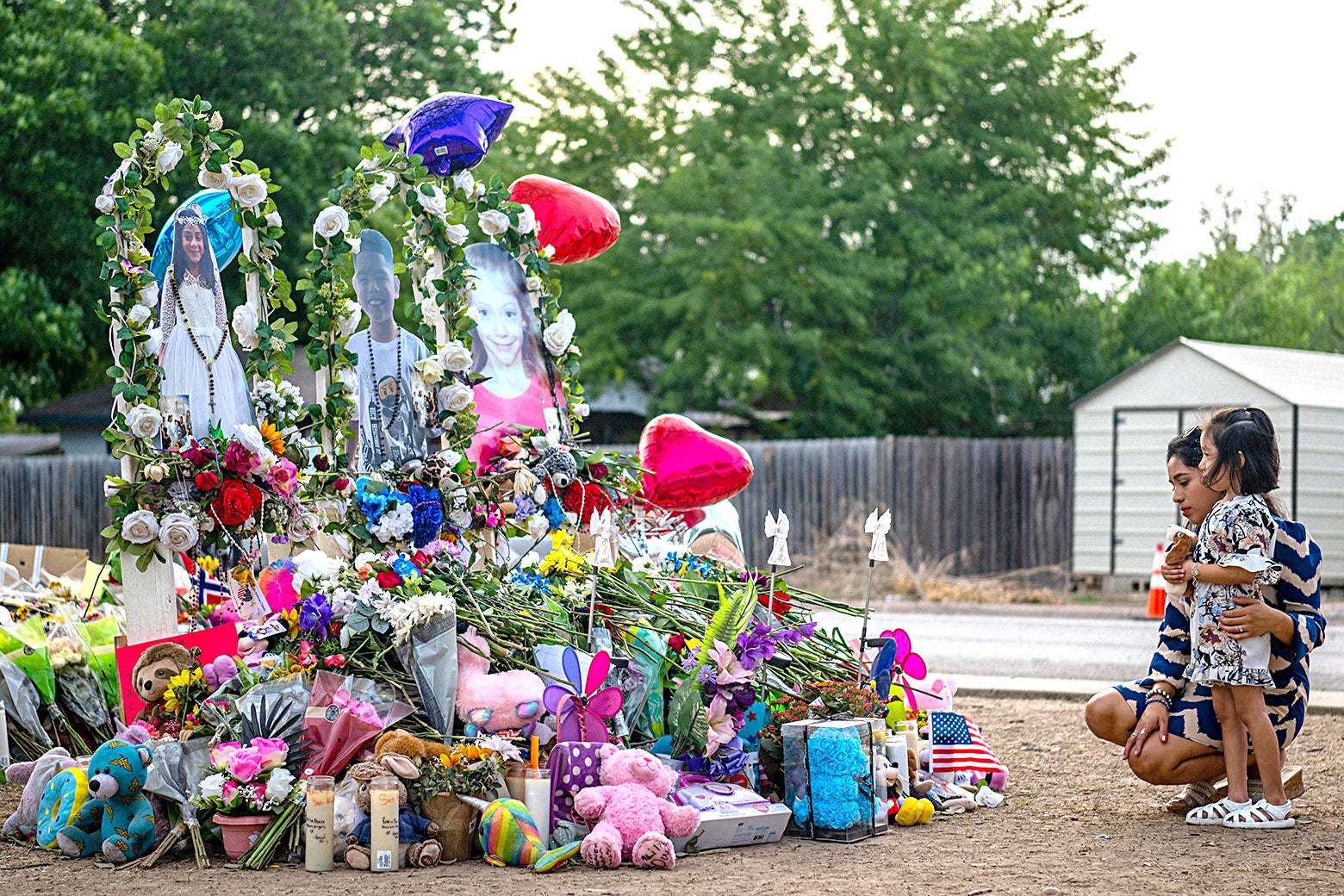 A woman crouches next to a young girl as they look at a memorial of flowers, stuffed animals, angels, candles, and photographs of the victims.