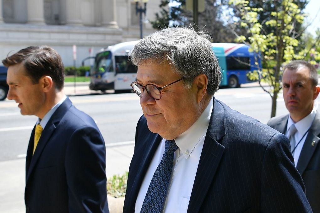Barr, wearing a suit, walks on a sidewalk with a half-smile on his face.