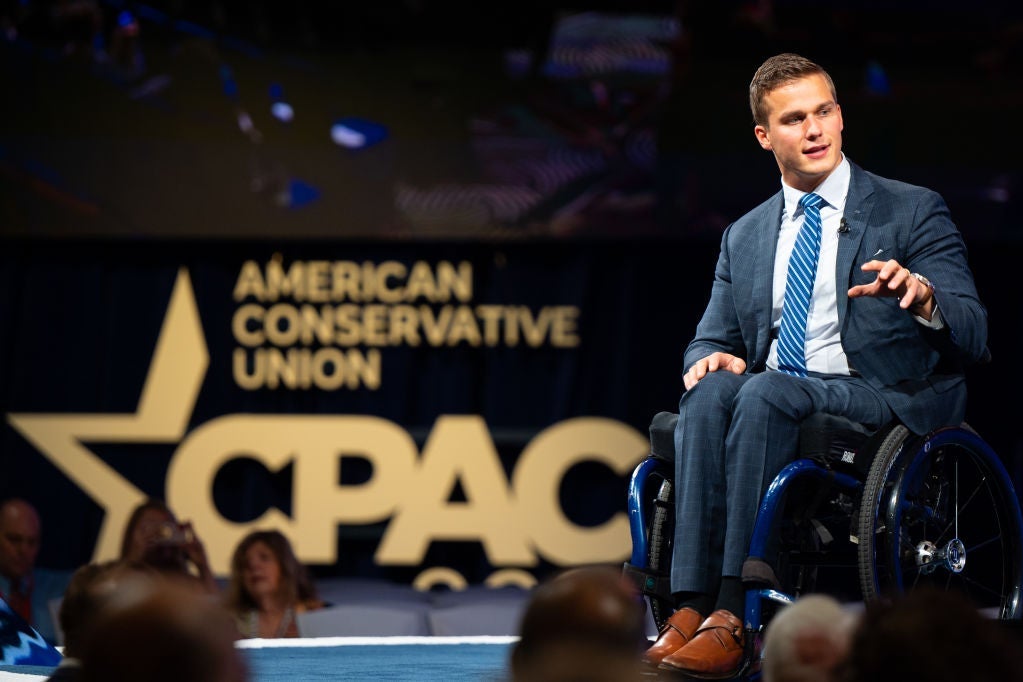 Cawthorn, wearing a blue suit and tie, gestures while speaking onstage in front of a CPAC backdrop.