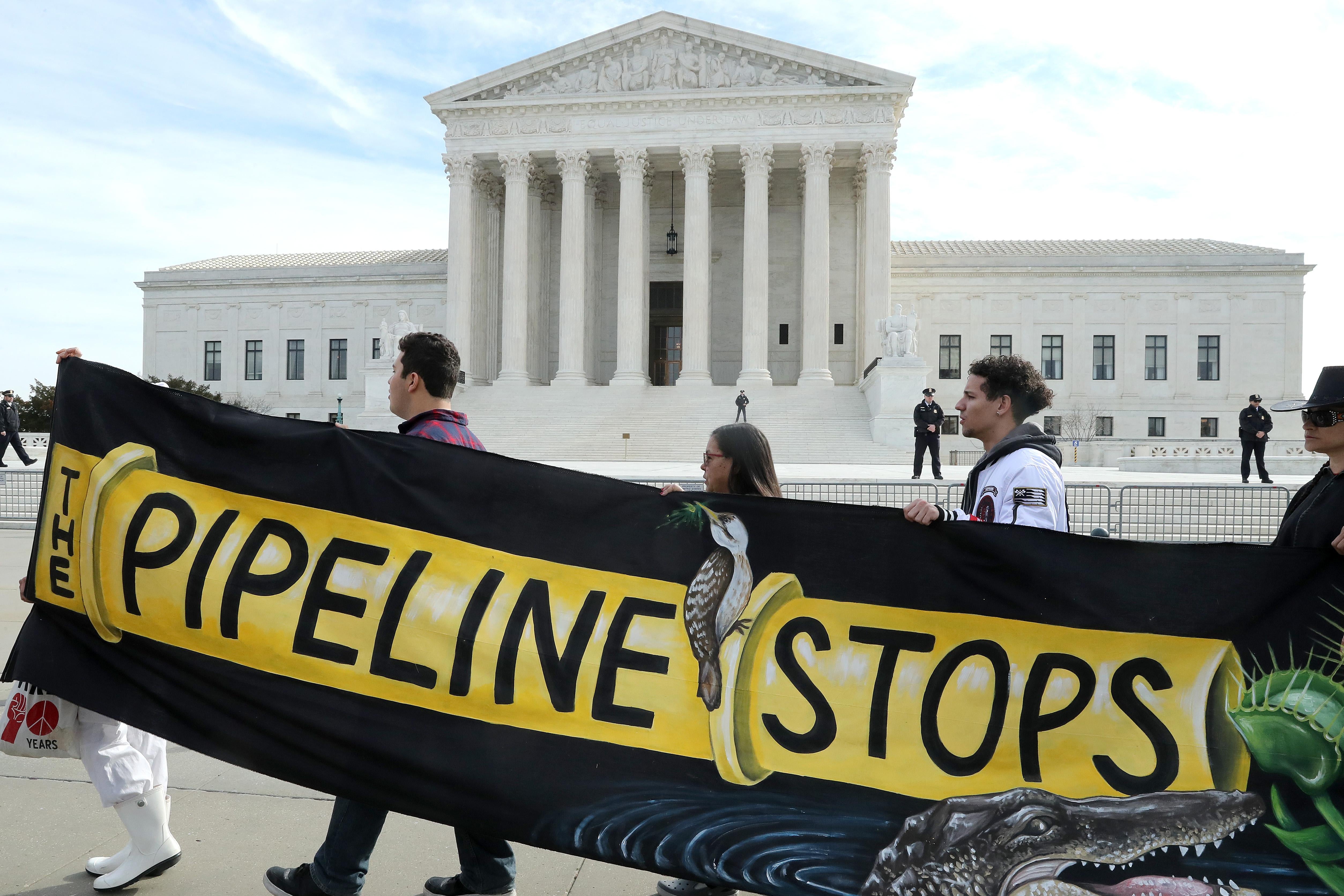 Protesters walk in front of the Supreme Court holding a banner with a picture of a pipeline on it saying "The Pipeline Stops."