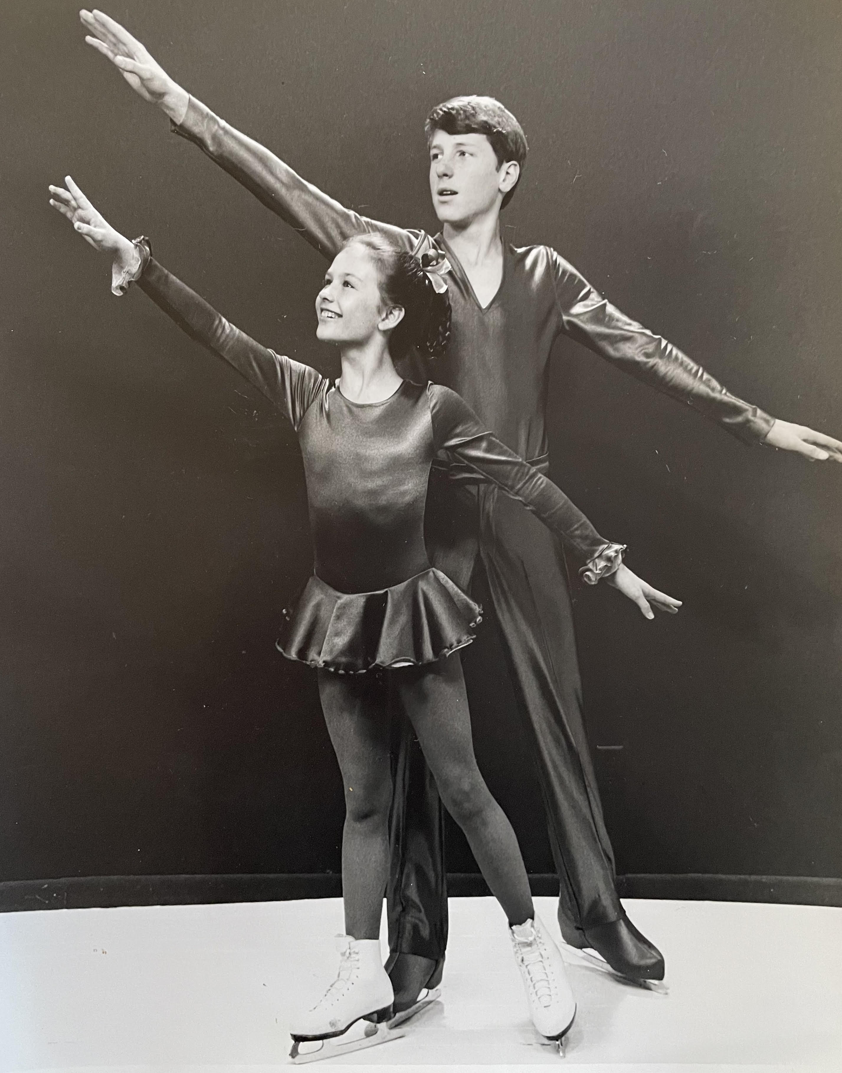 A girl and boy in ice dancing costumes and skates are posed each holding one arm up and one leg out. 