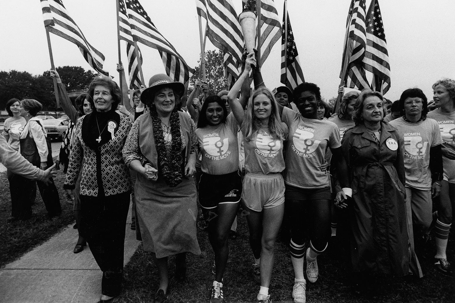 Women hold hands, smile, and carry torches and flags aloft.