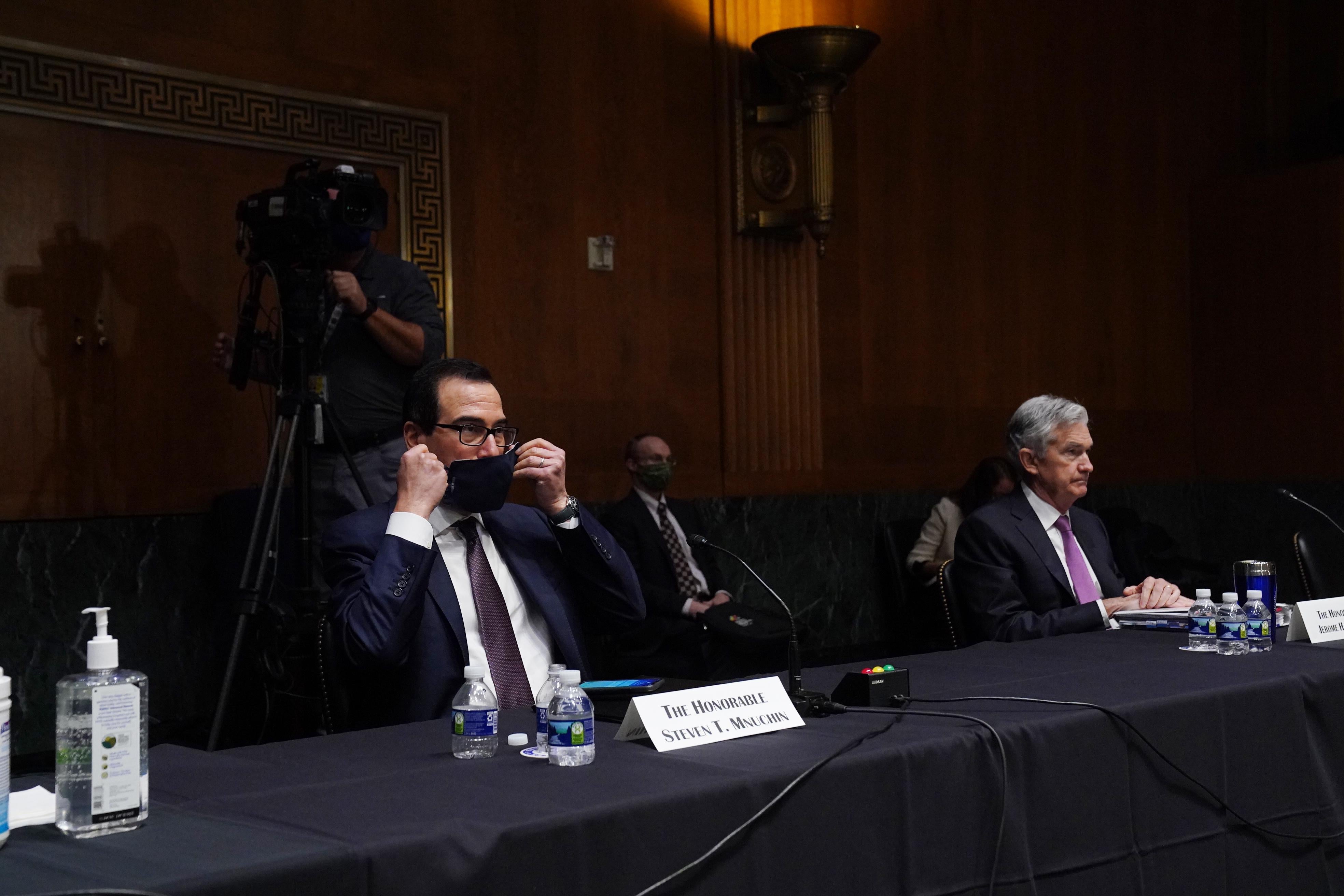 Mnuchin puts on a mask while sitting next to Jerome Powell at a long table in a Senate hearing room