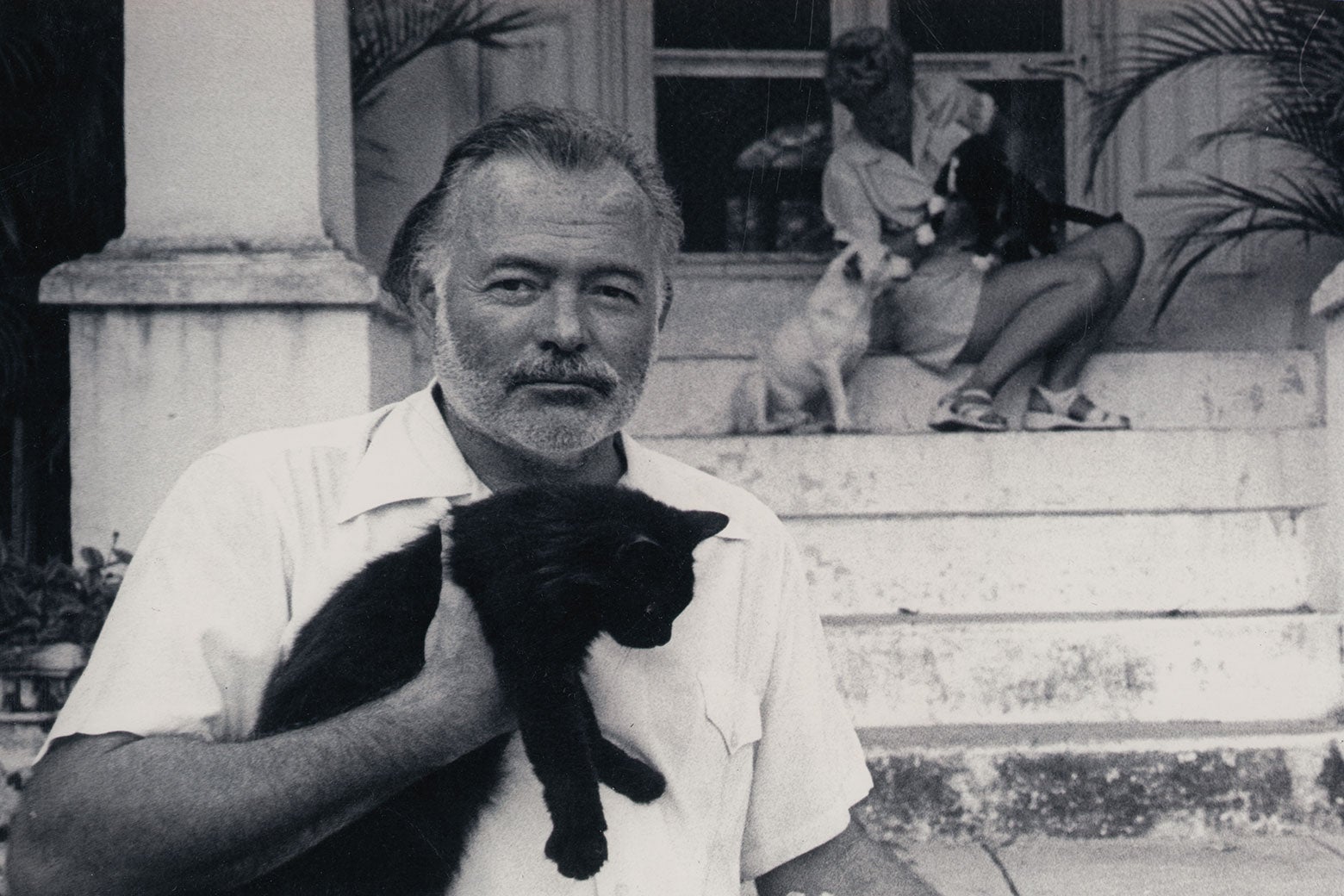Hemingway holds a cat while standing in front of a porch. A woman and a dog can be seen sitting on the steps behind him.