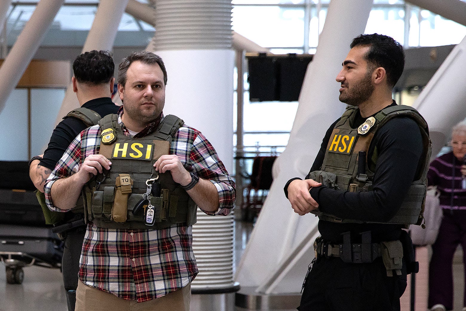 Two male ICE agents at the airpot, one in plaid button-up, looking sloppy. 