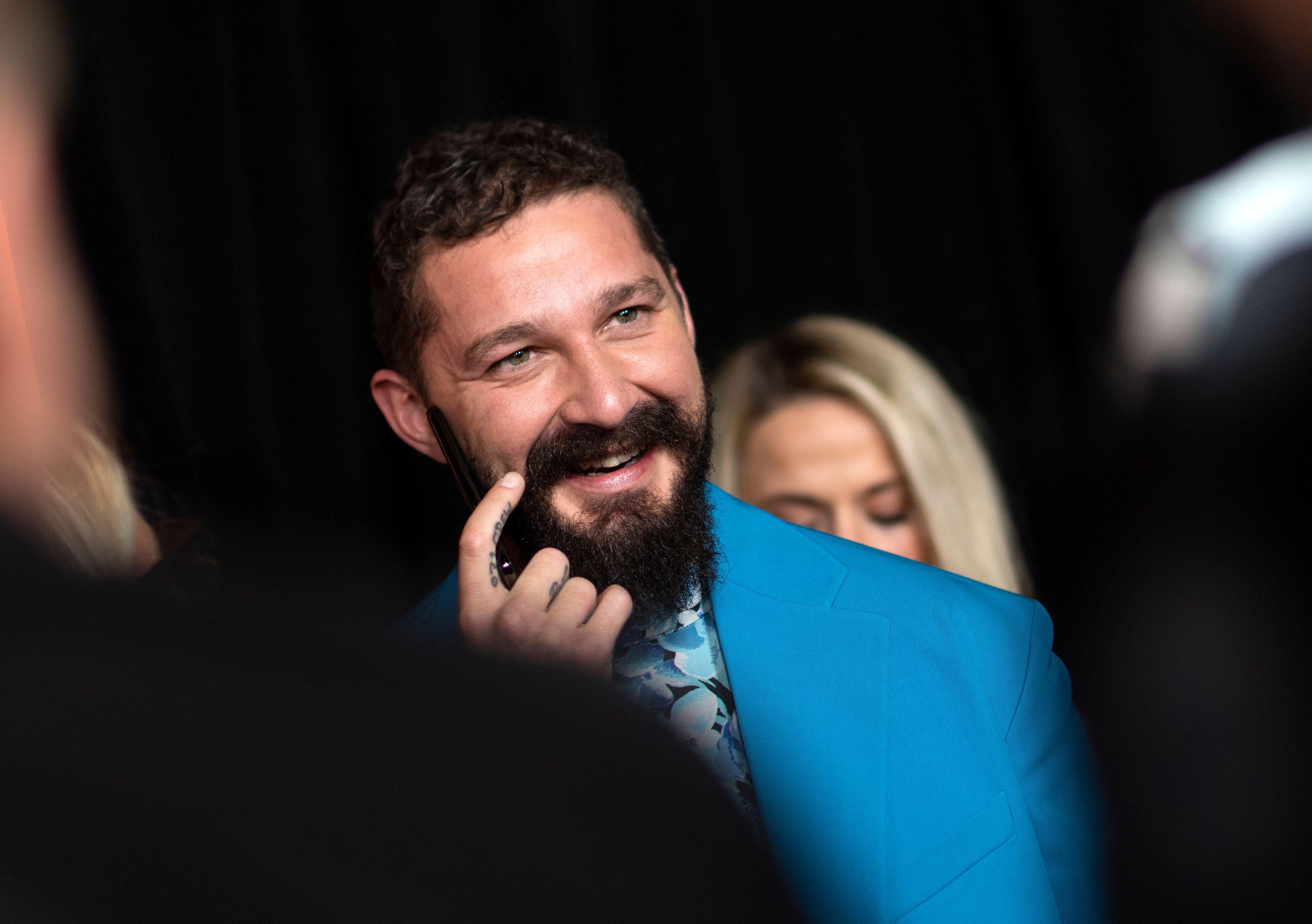 Shia LaBeouf, in a beard and blue suit, scratches his face and smiles.
