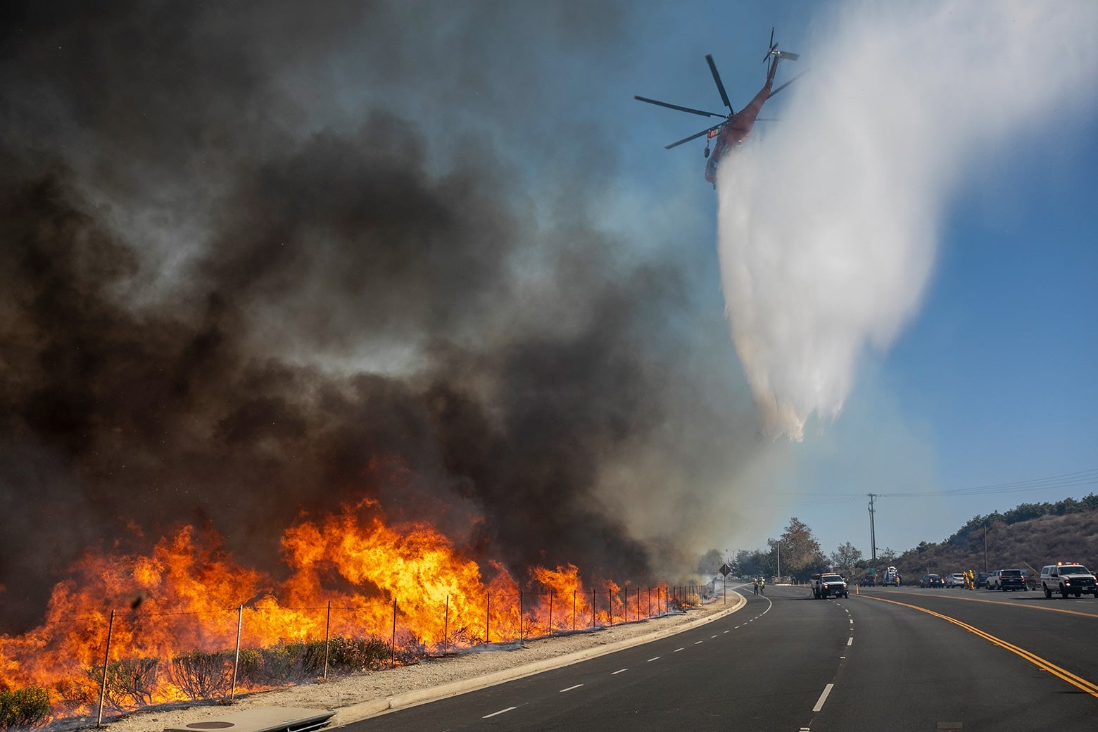 A helicopter drops water near a fire that is raging on the side of a road.