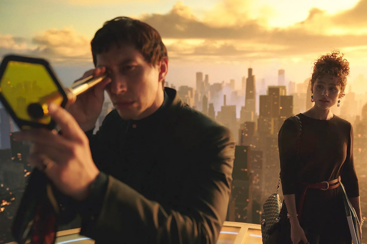 Driver, wearing a suit, looks through a tiny golden telescope at the top of a futuristic cityscape. Behind him, Emmanuel, looking elegant in a black dress, looks on.