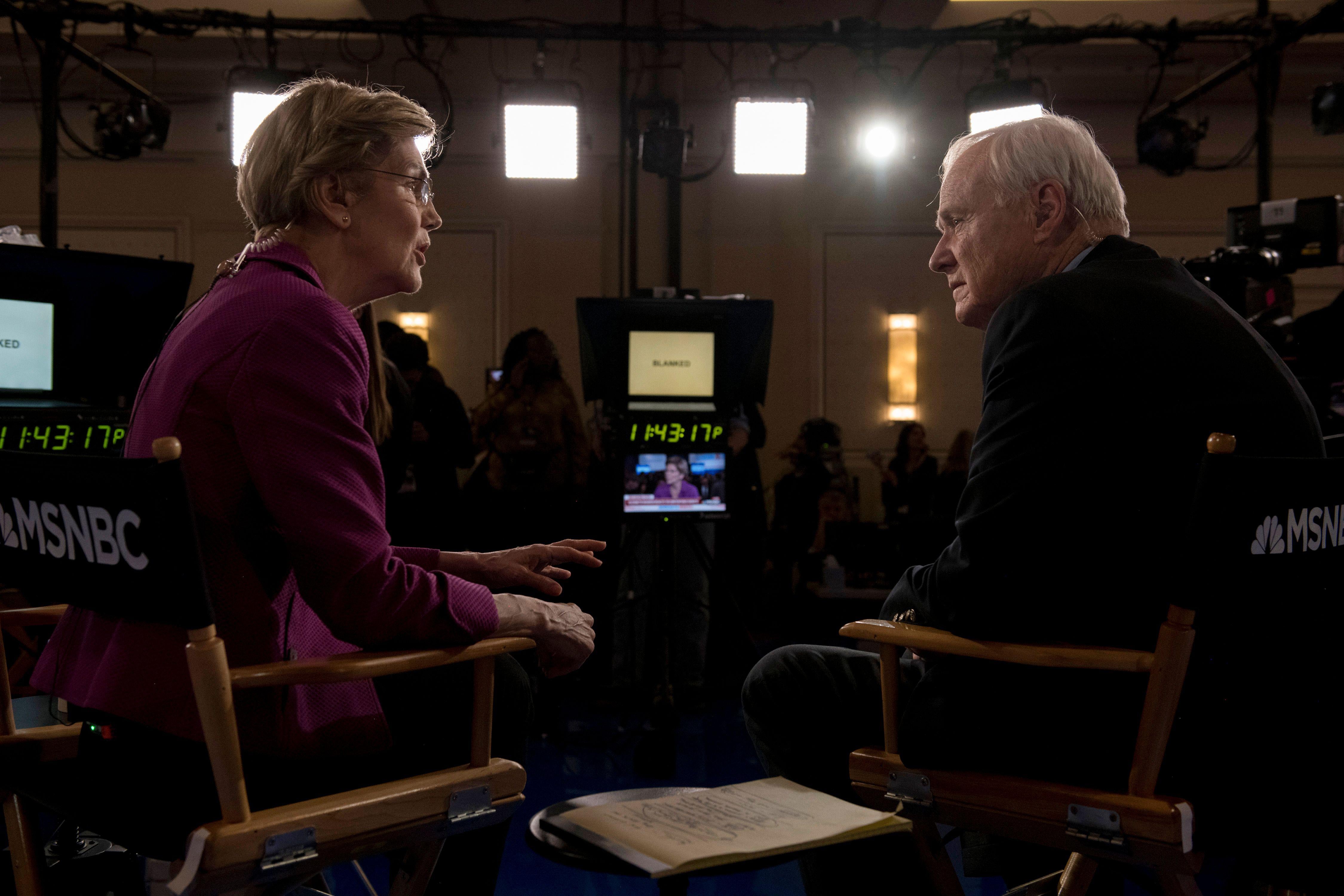 Elizabeth Warren and Chris Matthews speak together in the spin room after the South Carolina debate, sitting in folding chairs, cameras visible.