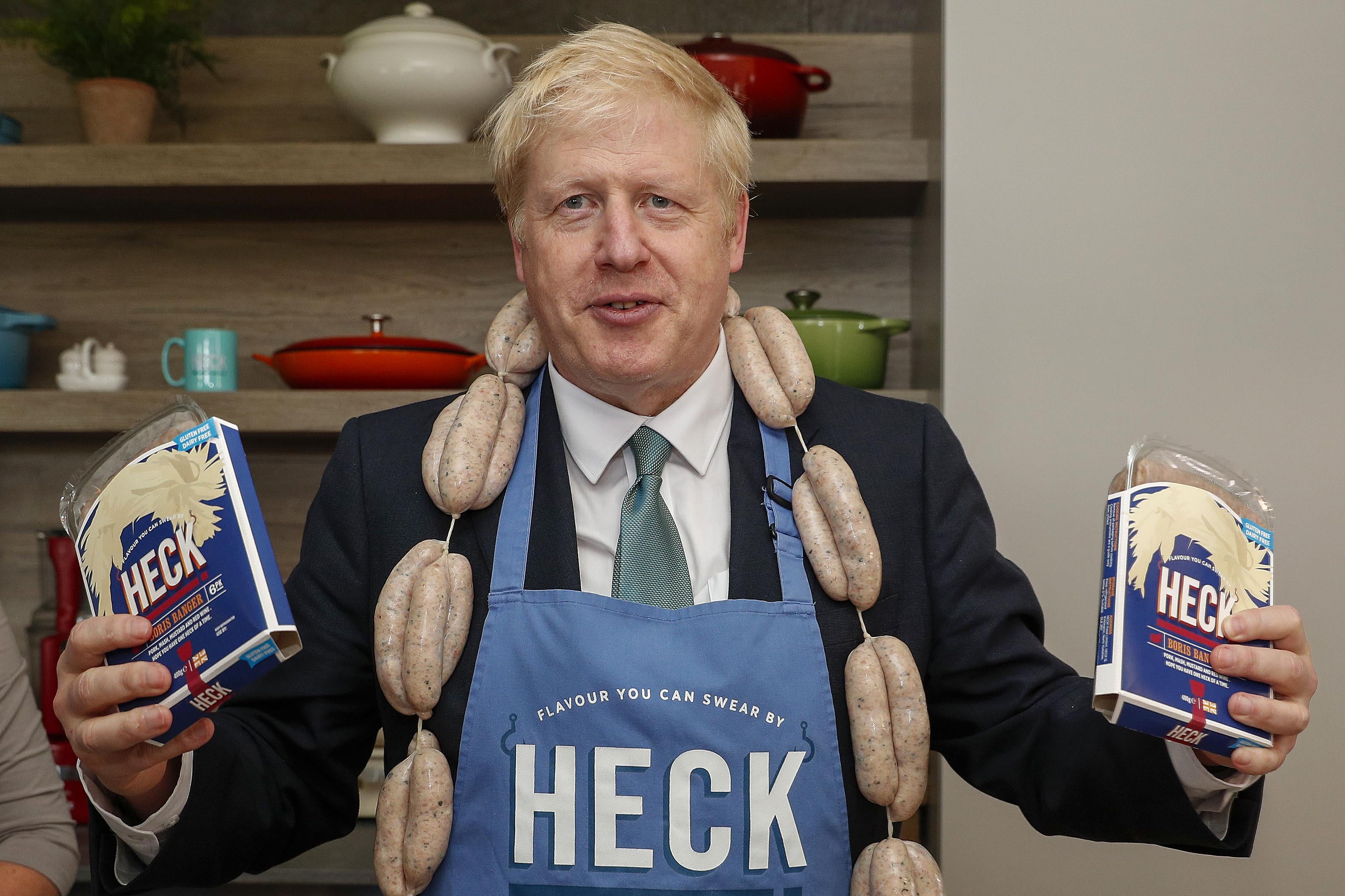 Boris Johnson holds a pack of  "Boris Bangers" sausages in each hand, wears a string of sausages around his neck like a scarf, and wears an apron that says "Heck" on it.