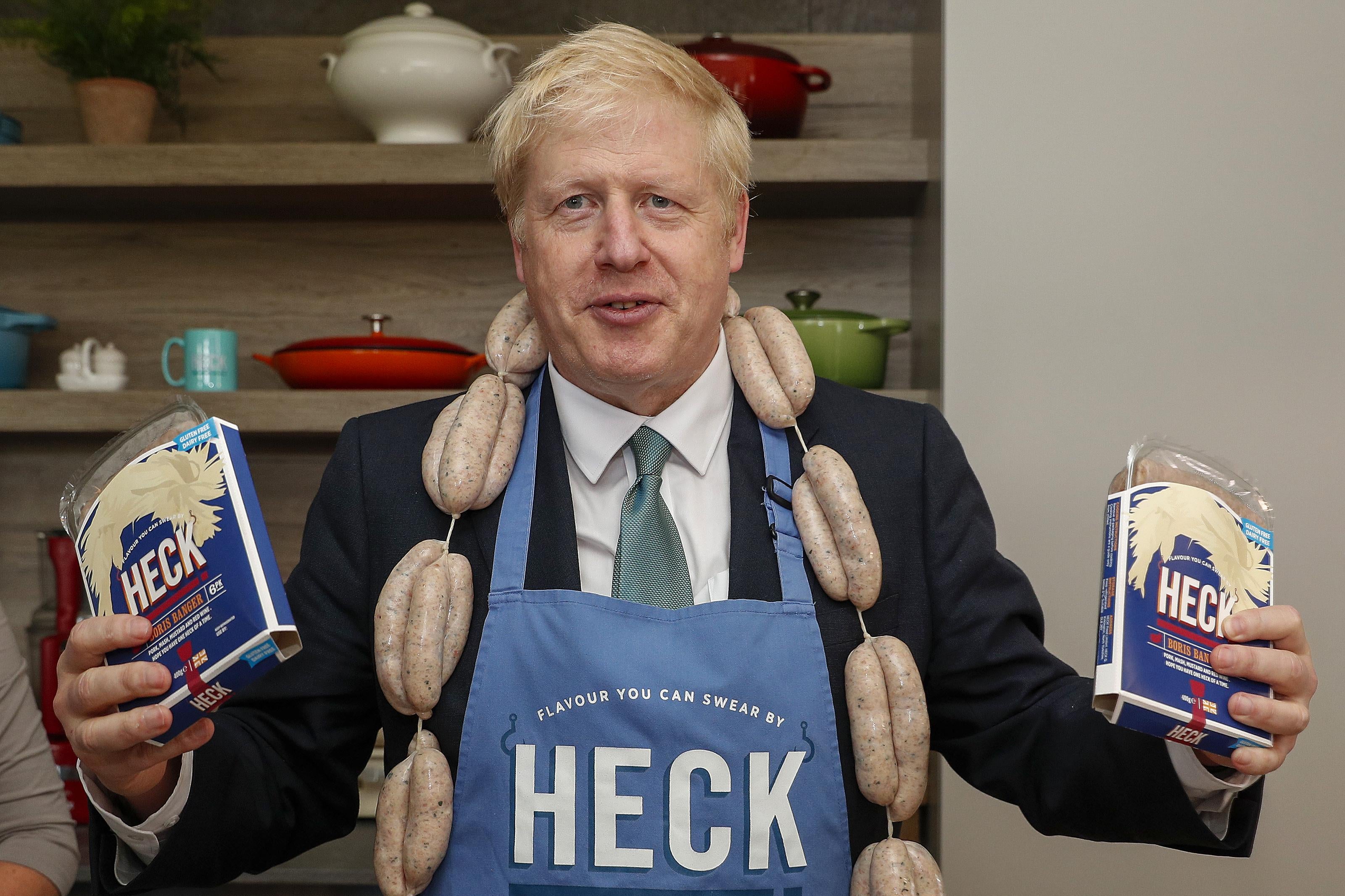 Boris Johnson holds a pack of "Boris Bangers" sausages in each hand, wears a string of sausages around his neck like a scarf, and wears an apron that says "Heck" on it.
