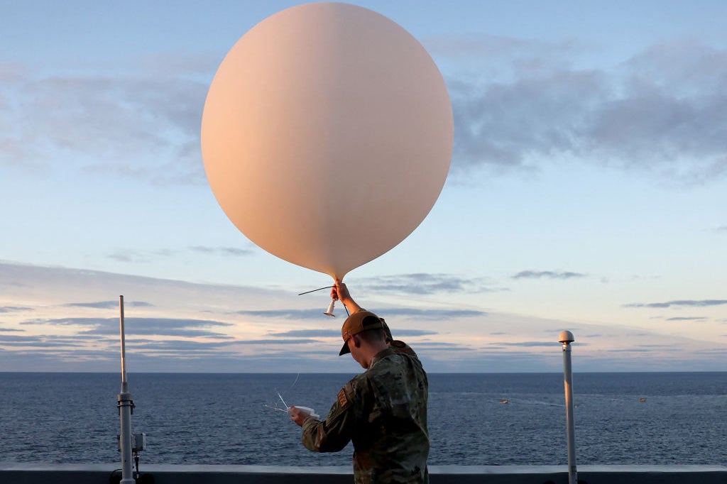 An airman in fatigues standing next to the rail of a ship holds up a white balloon that is about six feet in diameter.