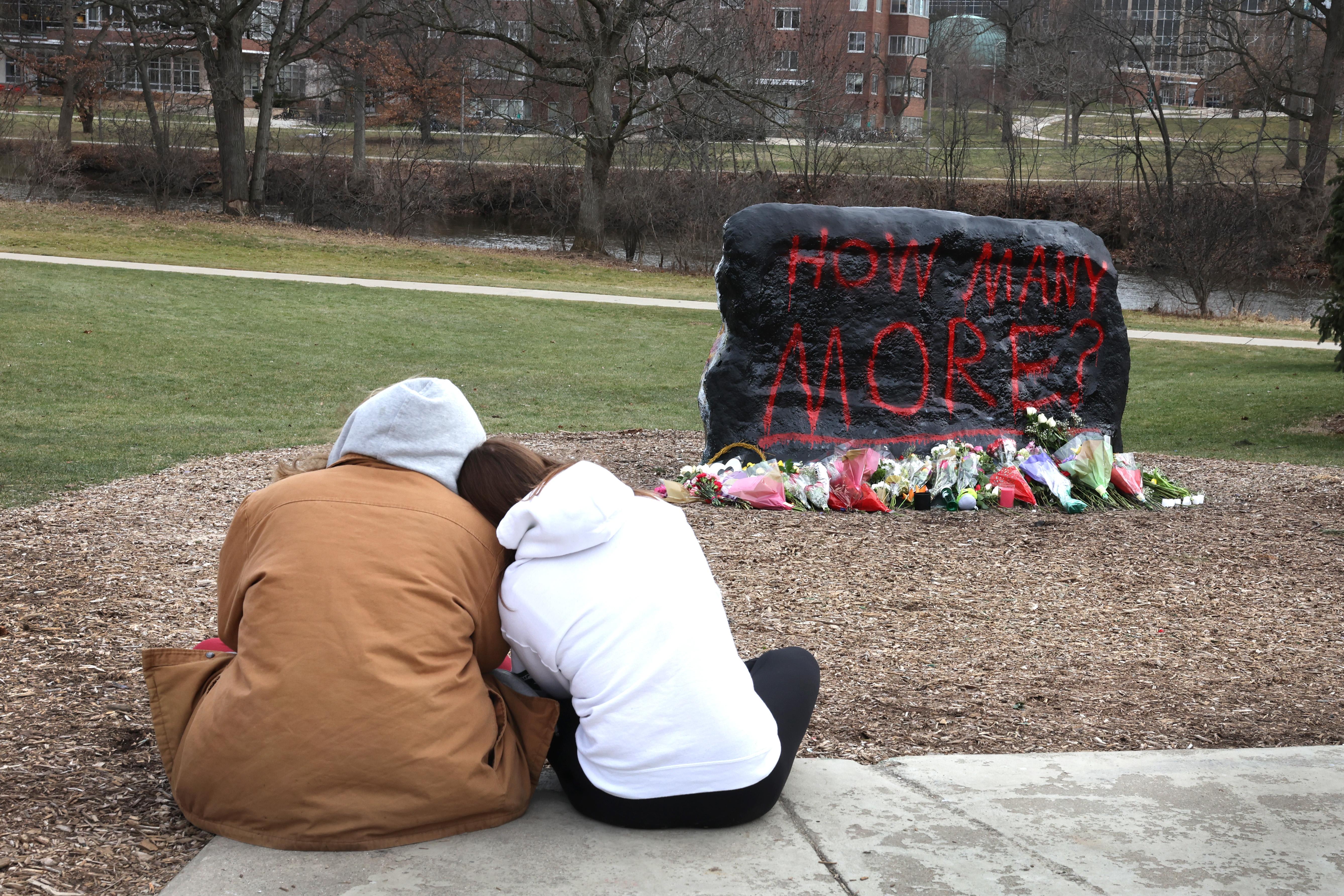 Two people, backs turned to the camera, huddle close to each in front of a rock on which the words "How Many More?" are painted.