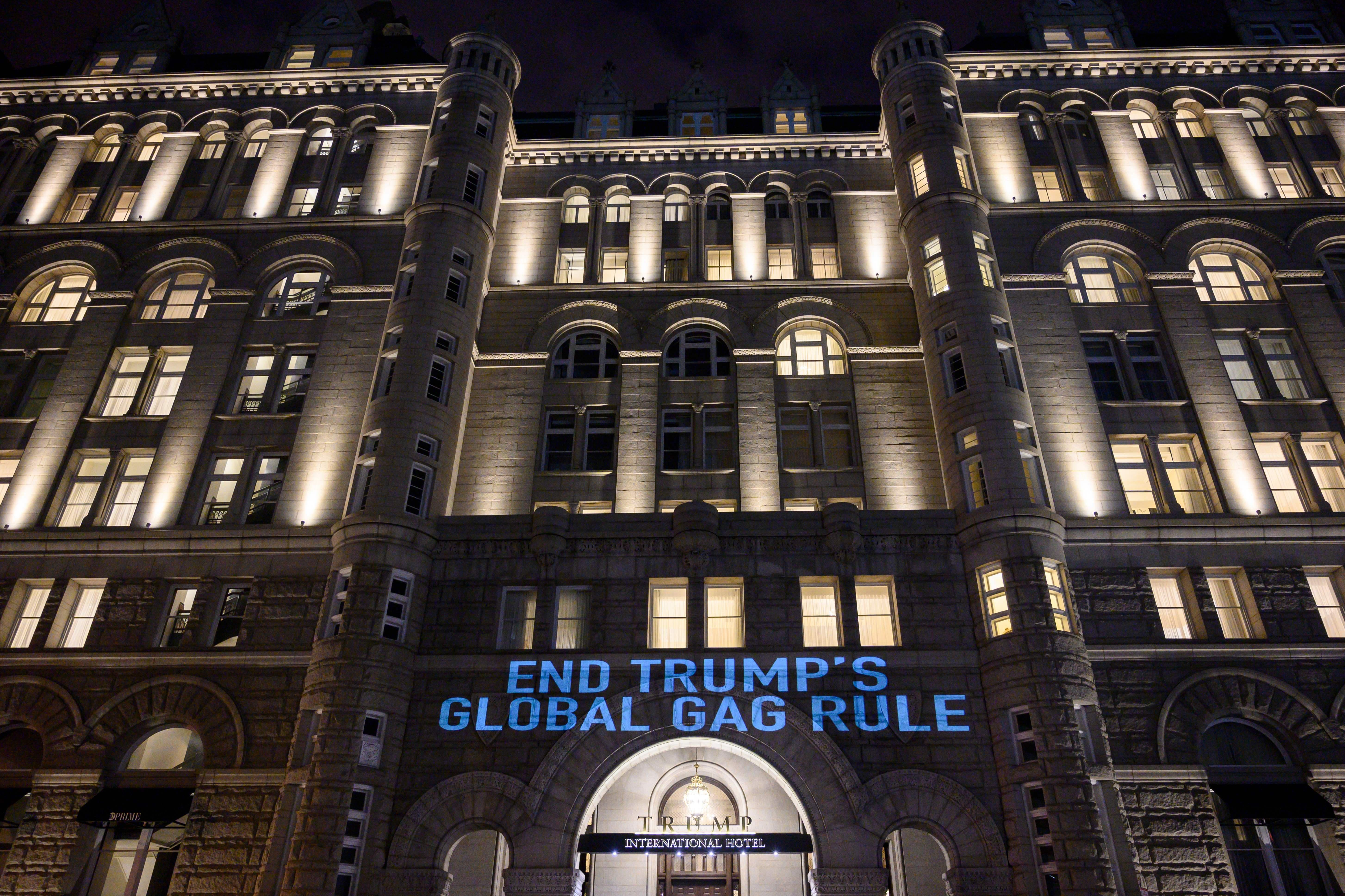 END TRUMP'S GLOBAL GAG RULE is projected in blue light on the front of dramatic-looking hotel at night.