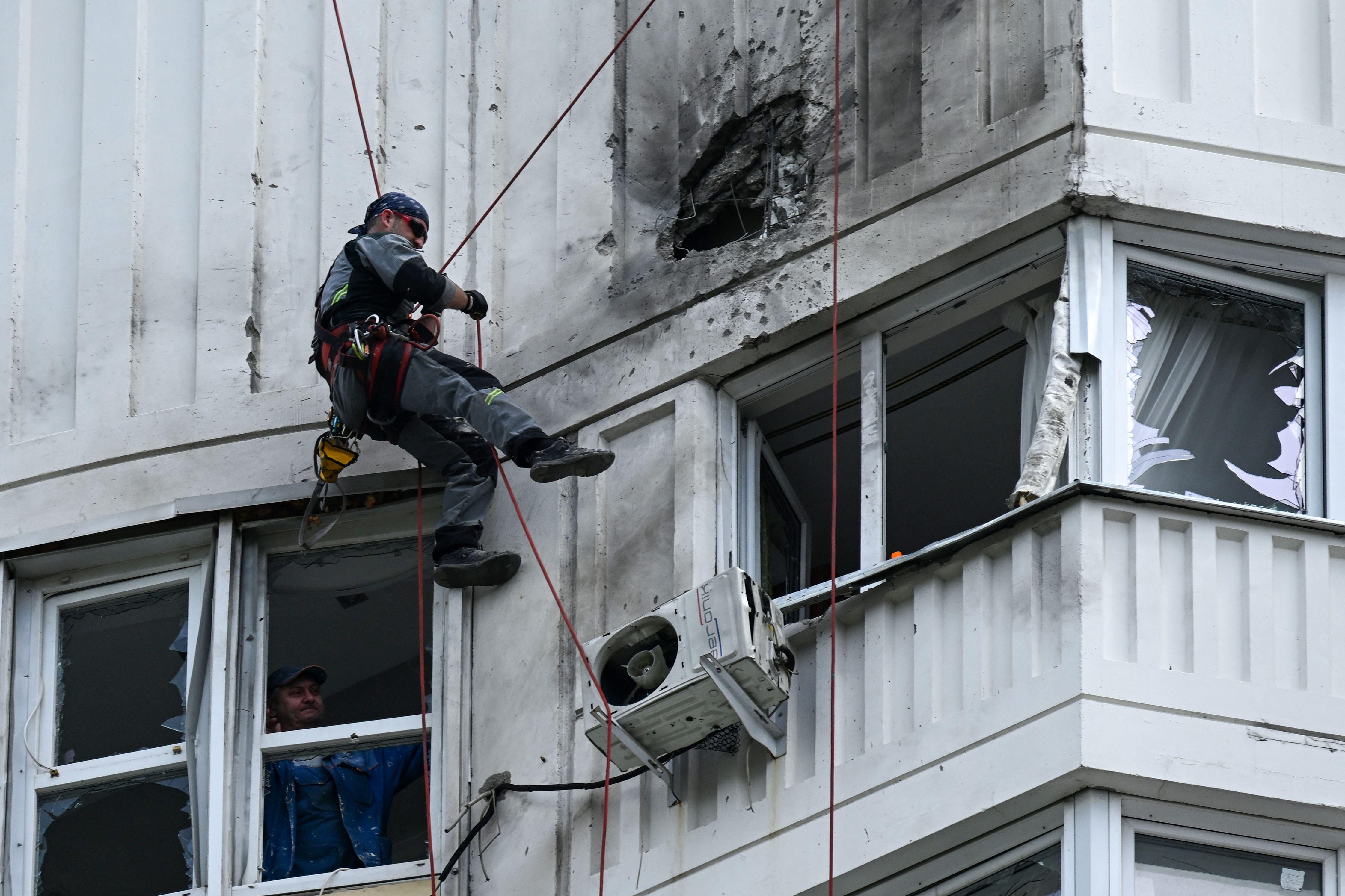 A person harnessed to the side of an apartment building inspects a hole blasted into its side. Broken windows are visible below.