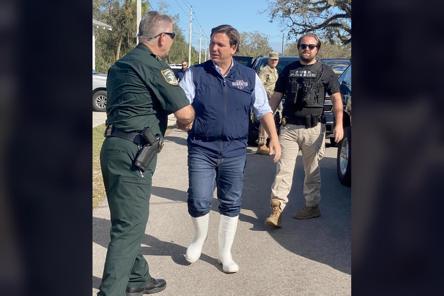DeSantis, wearing a blue vest and comically enormous white boots, shakes a law enforcement officer's hand.